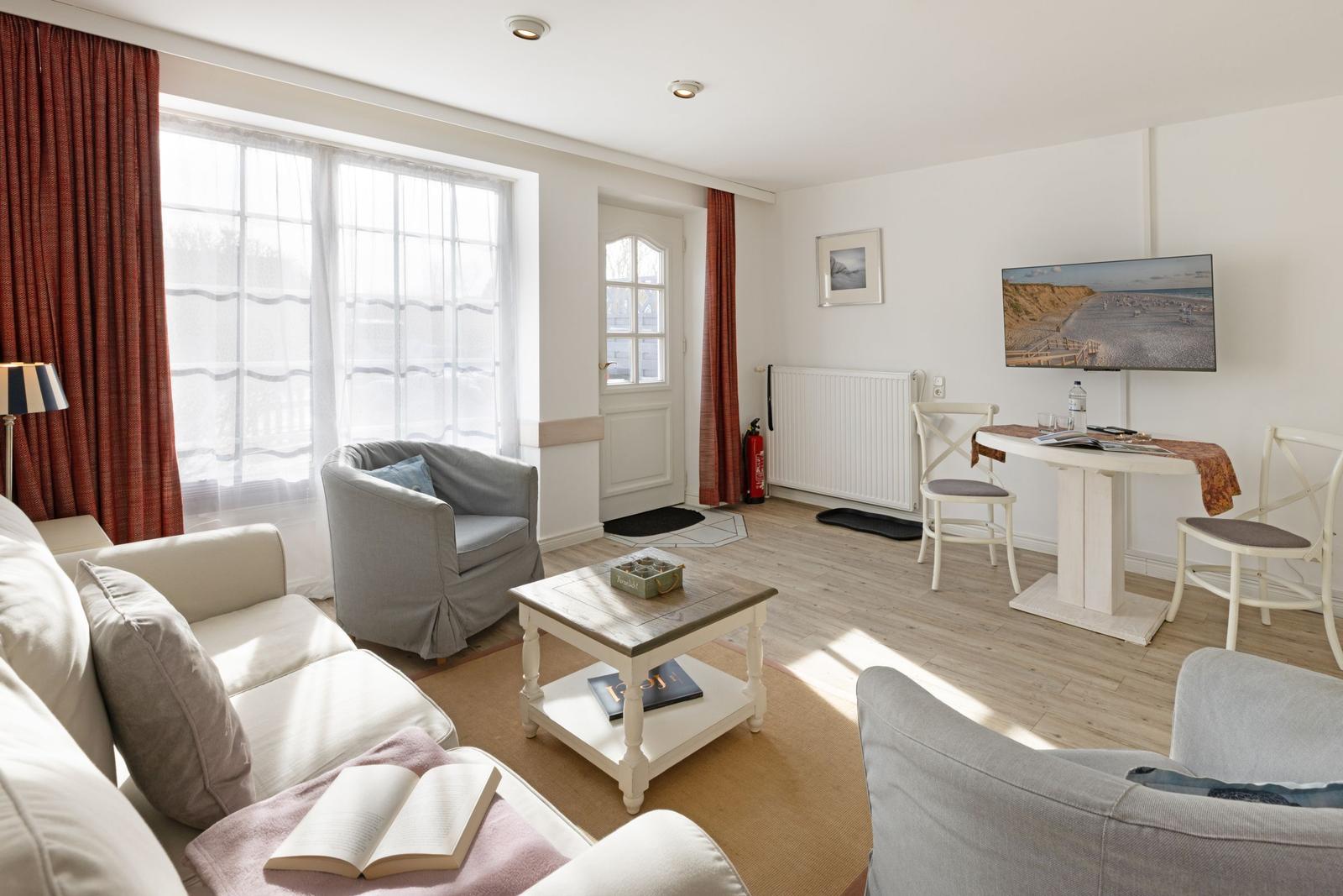 Wohnzimmer mit Sofa, Sessel, Esstisch und Fernseher. Fenster mit Vorhängen und Holzboden.