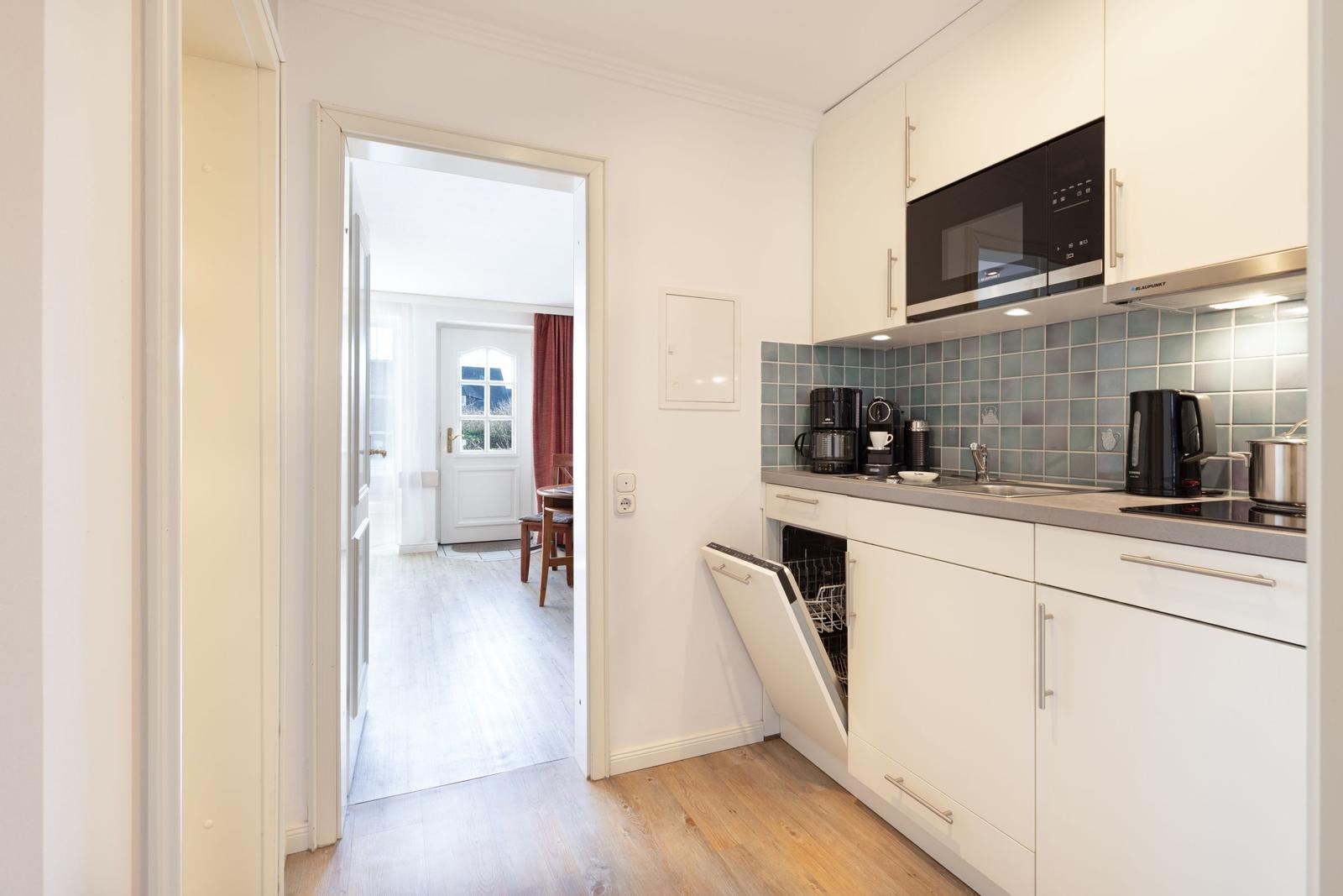 Kitchen with white cabinets, sink, and open dishwasher. Corridor to living area with wooden floor.