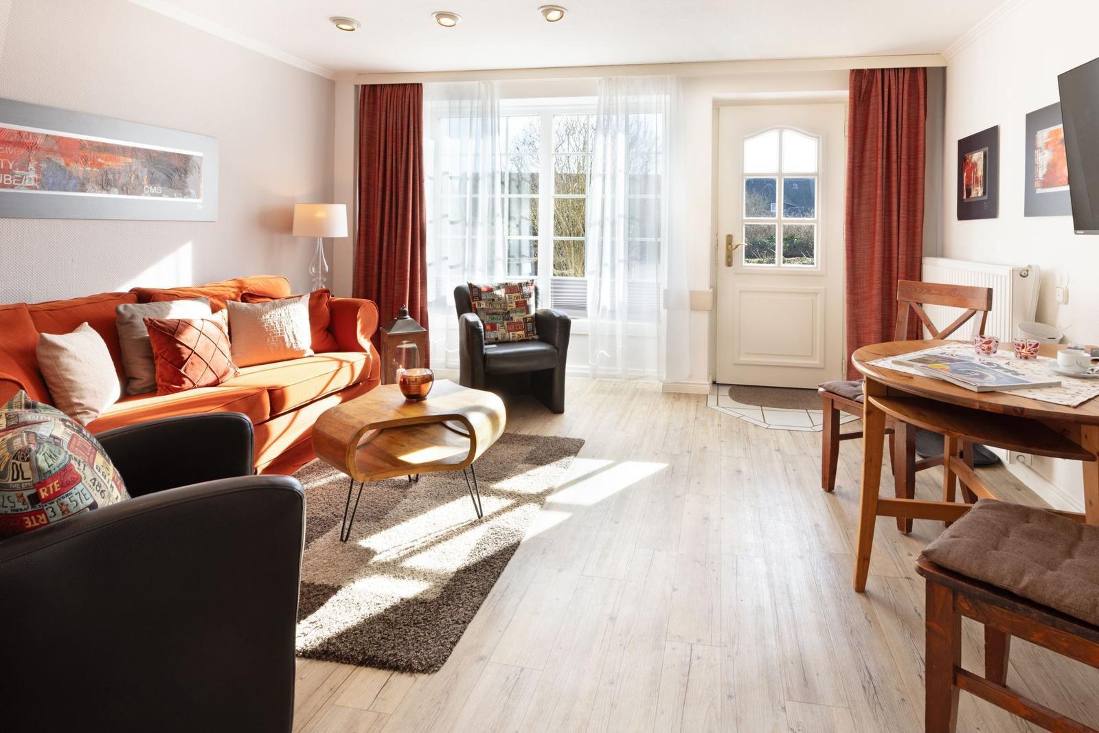 Living area with sofa, armchairs, and dining table. Wooden floor, windows, and door.