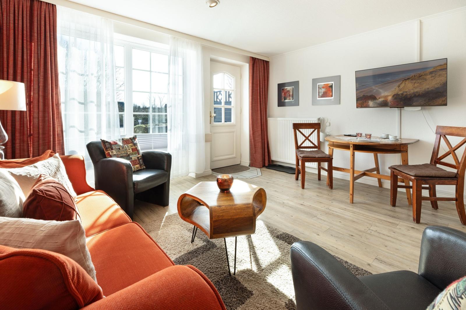 Living room with sofa, armchairs, table, and TV. Windows with curtains and door.