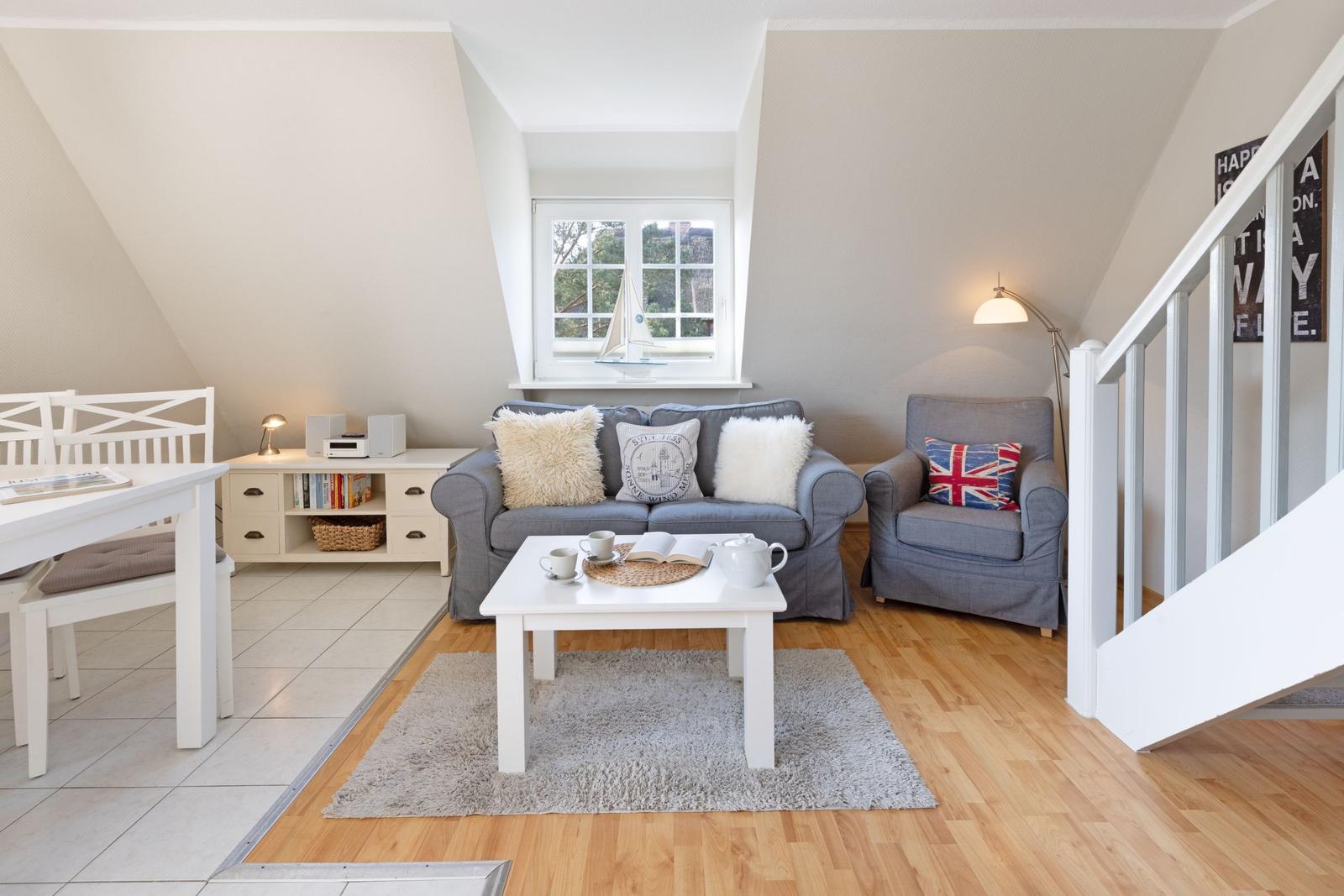 Wohnbereich mit grauem Sofa, weißem Tisch und Holzboden. Fenster mit Segelbootmodell.