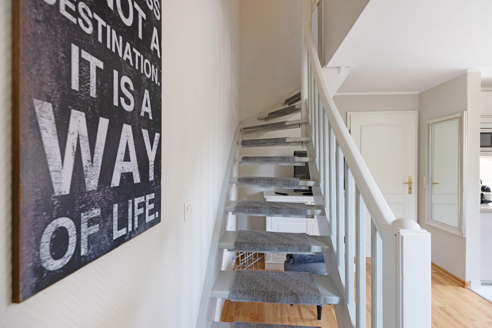 Treppenhaus mit weißen Geländern und grauen Teppichstufen. Wandbild mit Motivtext.