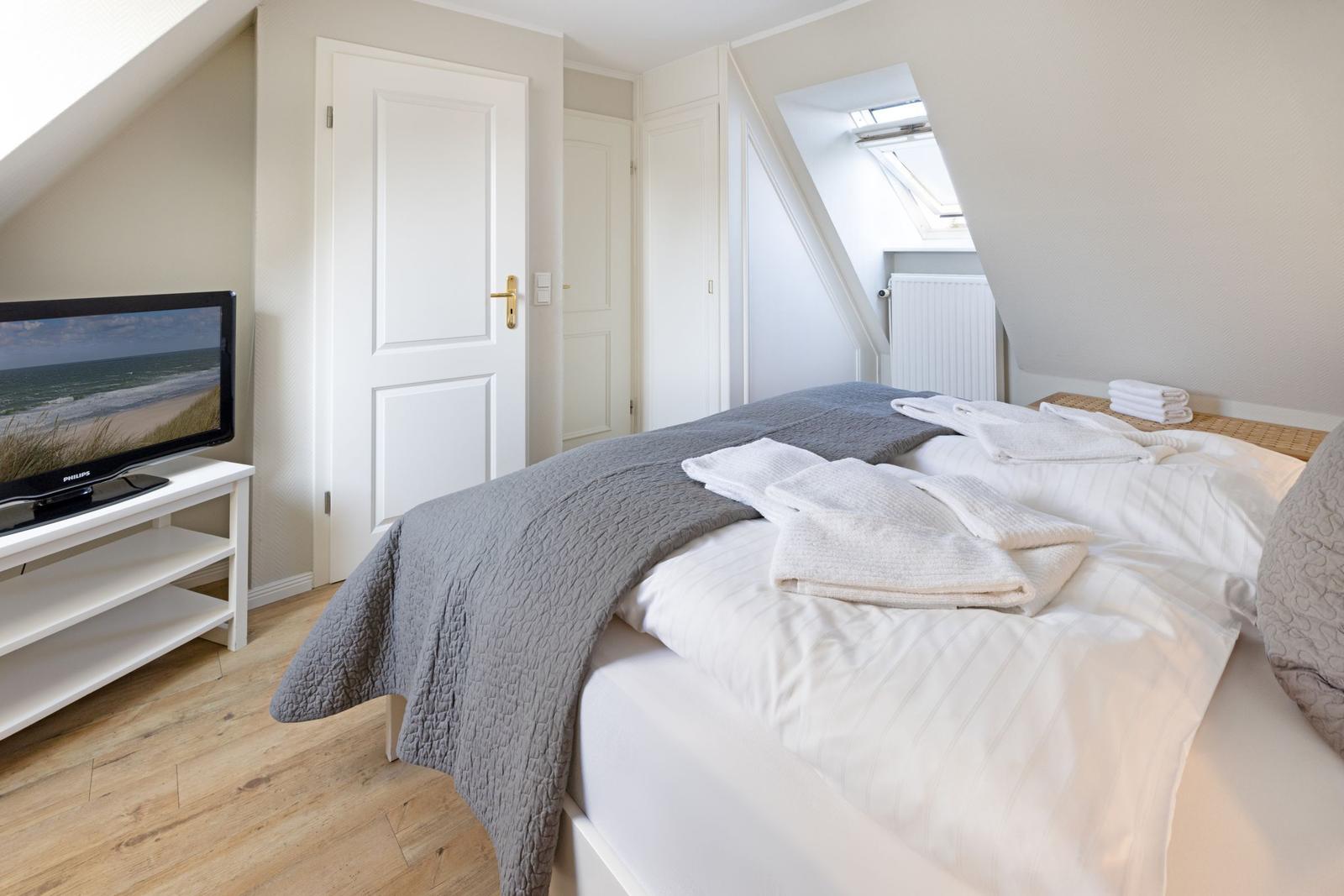 Schlafzimmer mit Dachschrägen, Bett mit weißen Laken und grauer Decke, Fernseher auf weißem Tisch.