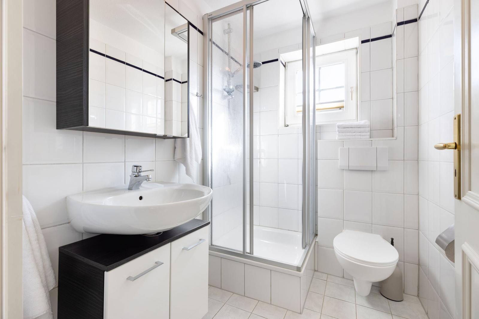 Badezimmer mit Dusche, Waschbecken und Toilette. Weiße Fliesen und schwarz-weiße Möbel.