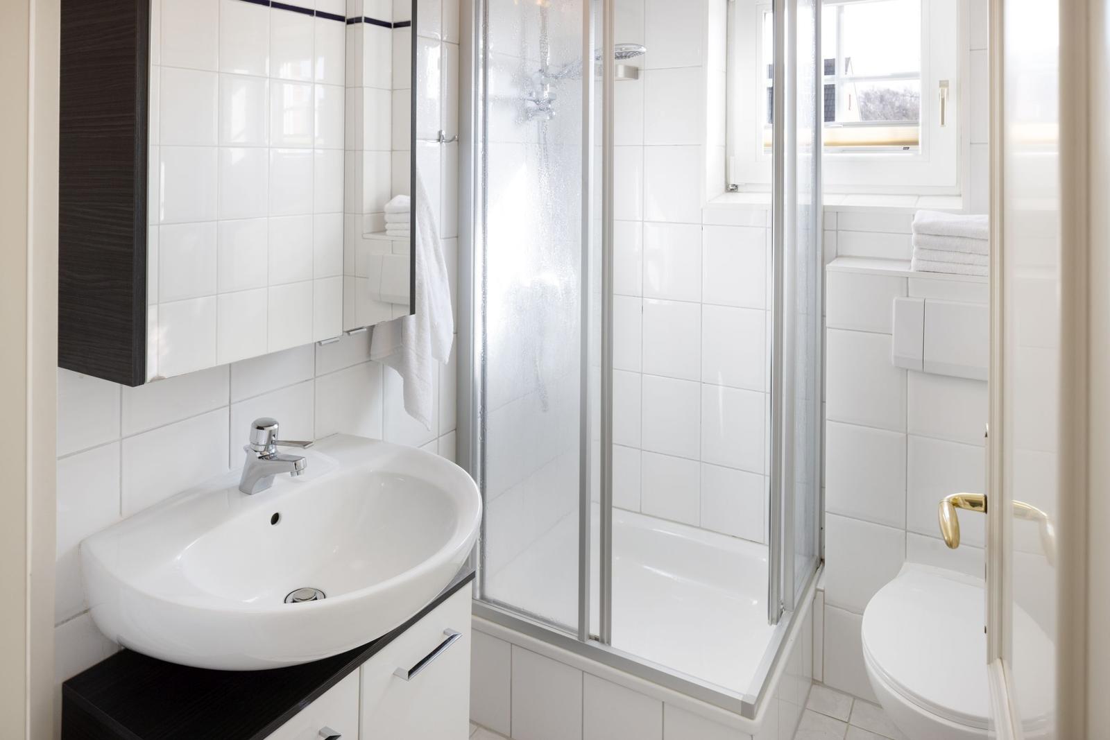 Badezimmer mit Dusche, Waschbecken und Toilette. Weiße Fliesen und schwarze Schrankfront.