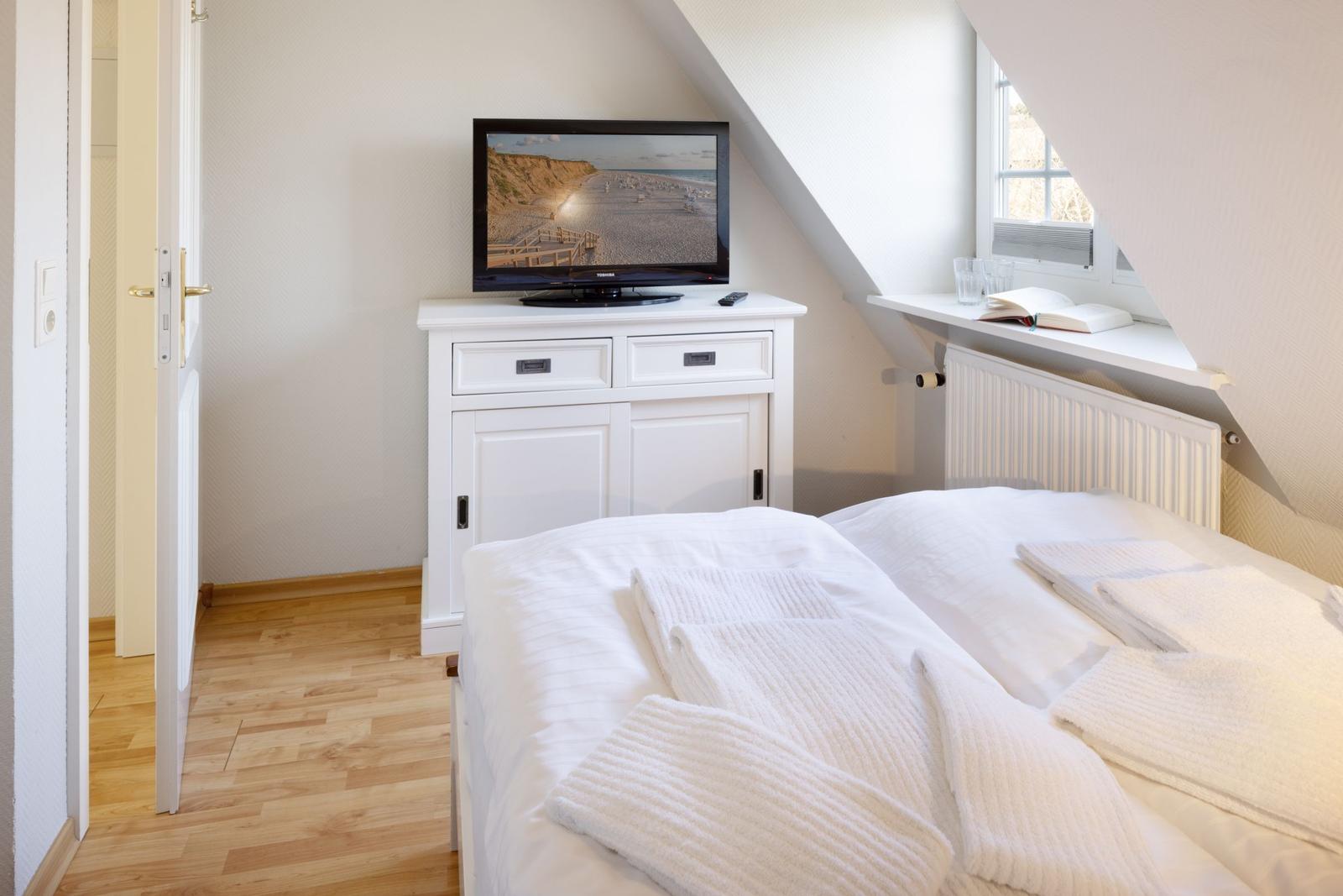 Schlafzimmer mit Bett, Fernseher und Fenster. Holzfußboden und weiß eingerichtet.