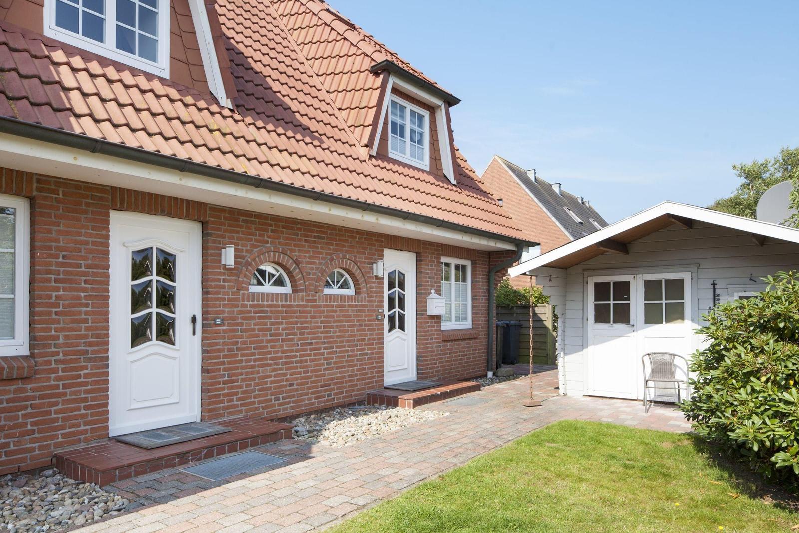Haus mit rotem Ziegeldach, Backsteinfassade und weißer Tür. Garten mit Rasen und Pflanzen.