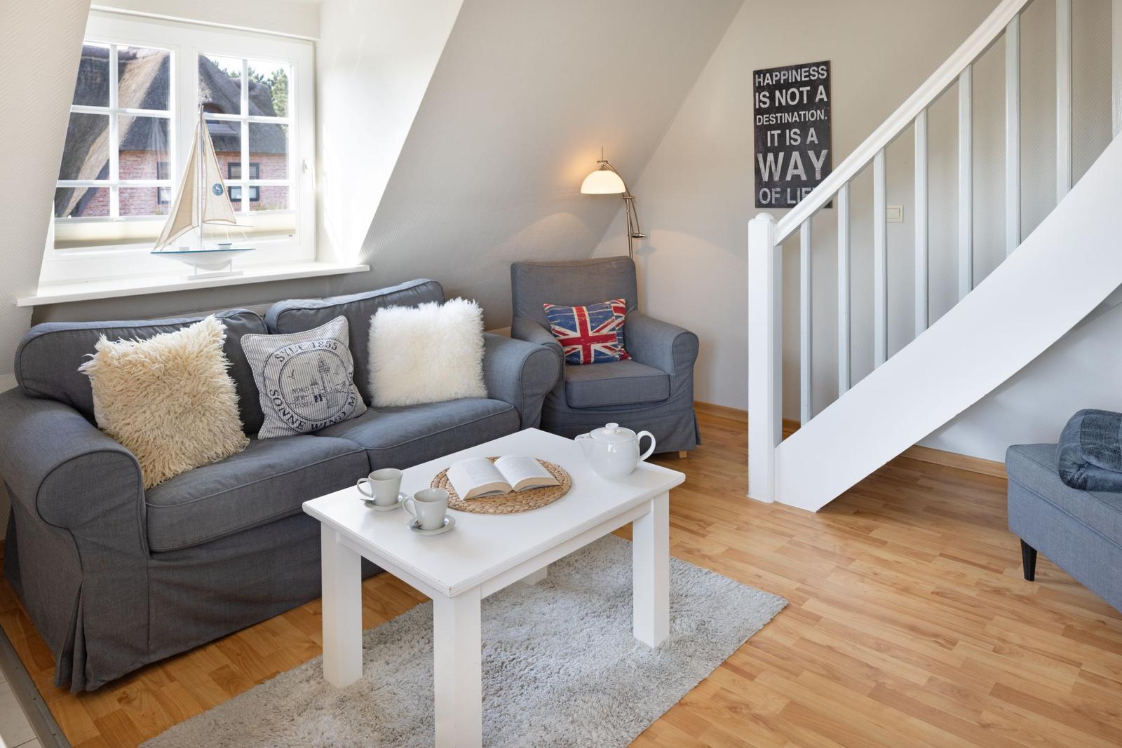 Wohnzimmer mit grauem Sofa, weißen Kissen und Holzboden. An der Wand hängt ein Poster.