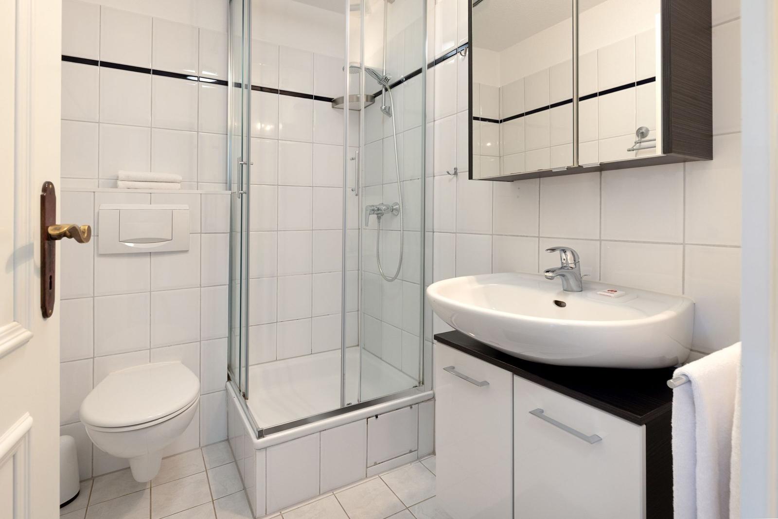 Badezimmer mit Dusche, Waschbecken und Toilette. Weiße Fliesen und dunkler Schrank.