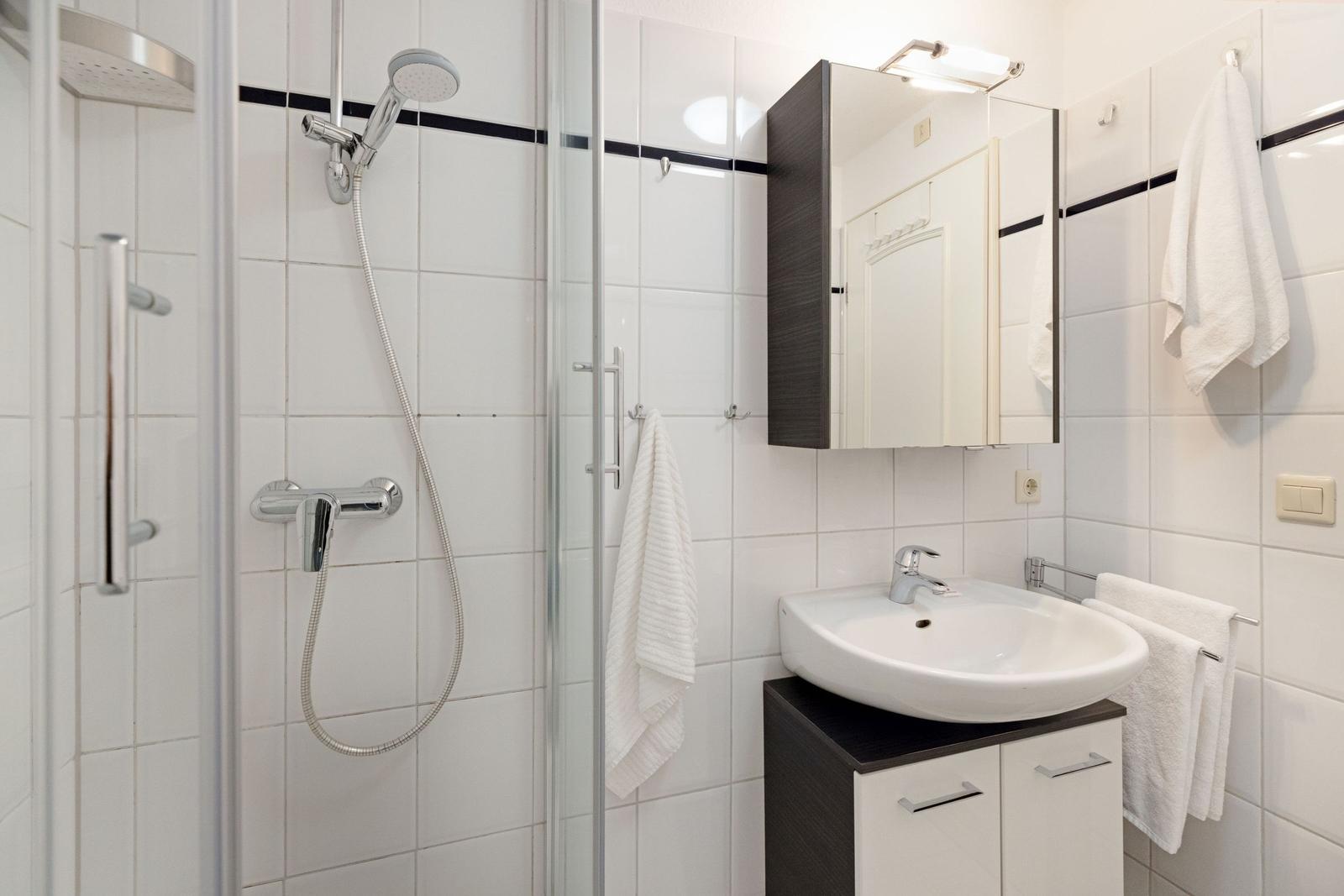 Badezimmer mit Dusche, Waschbecken und Spiegelschrank. Weiße Fliesen und Handtücher.