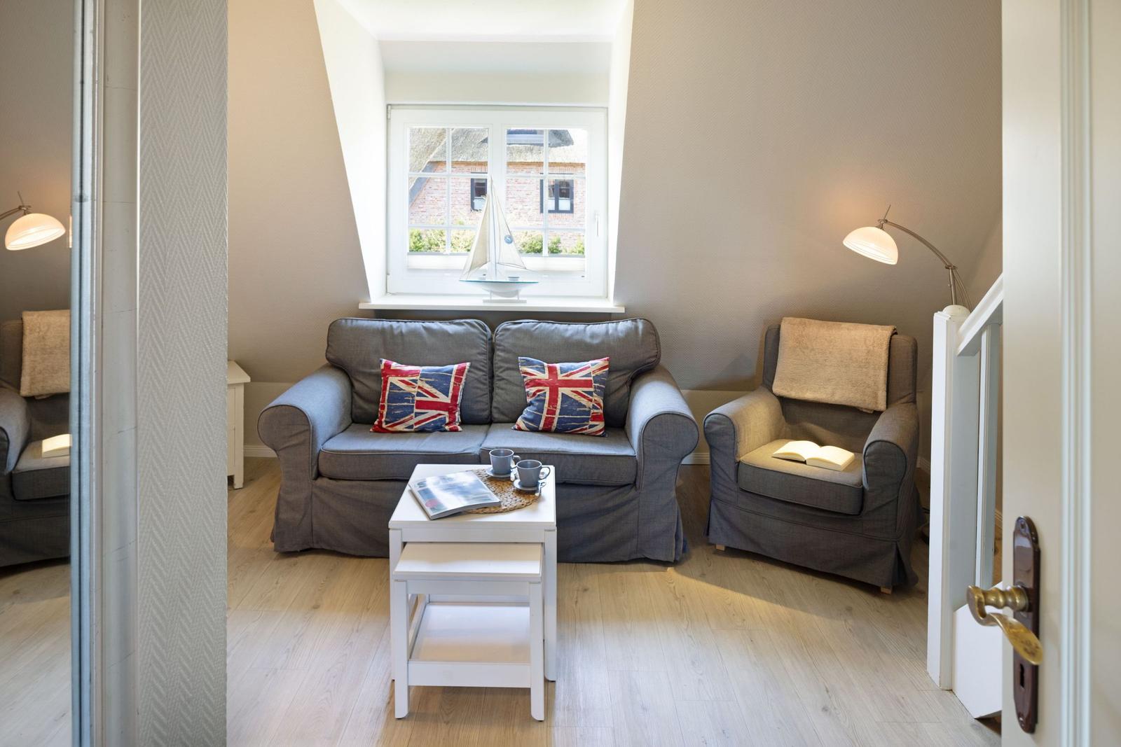 Wohnzimmer mit Sofa, Sessel und Fenster. Auf dem Sofa liegen Kissen mit Flaggenmuster.