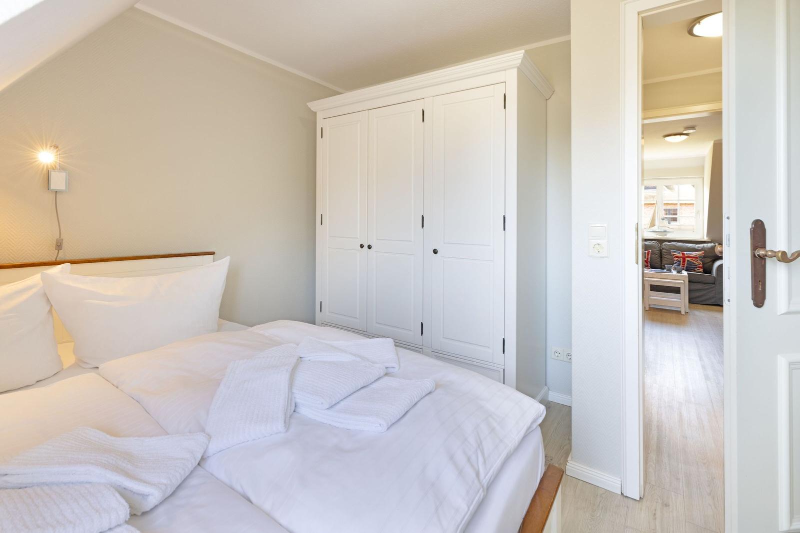 Schlafzimmer mit weißem Bett, Kleiderschrank und Blick in Wohnzimmer.