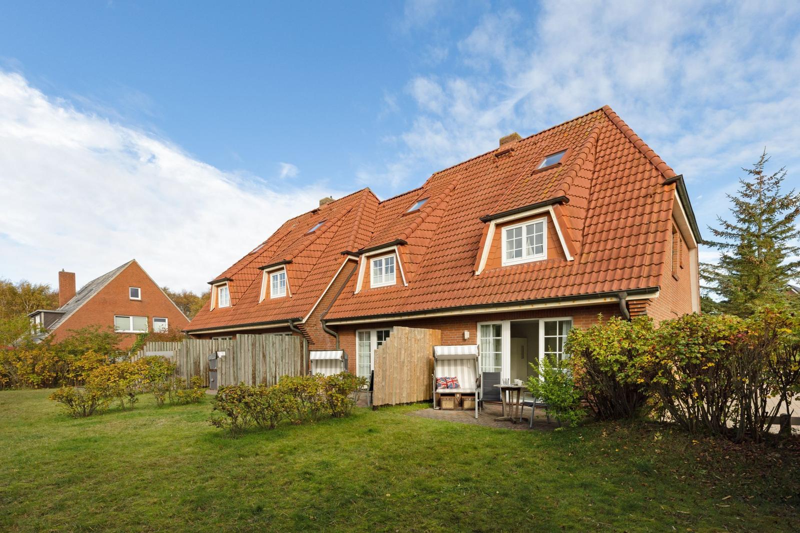A house with a red tiled roof, garden, and terrace with chair and table.