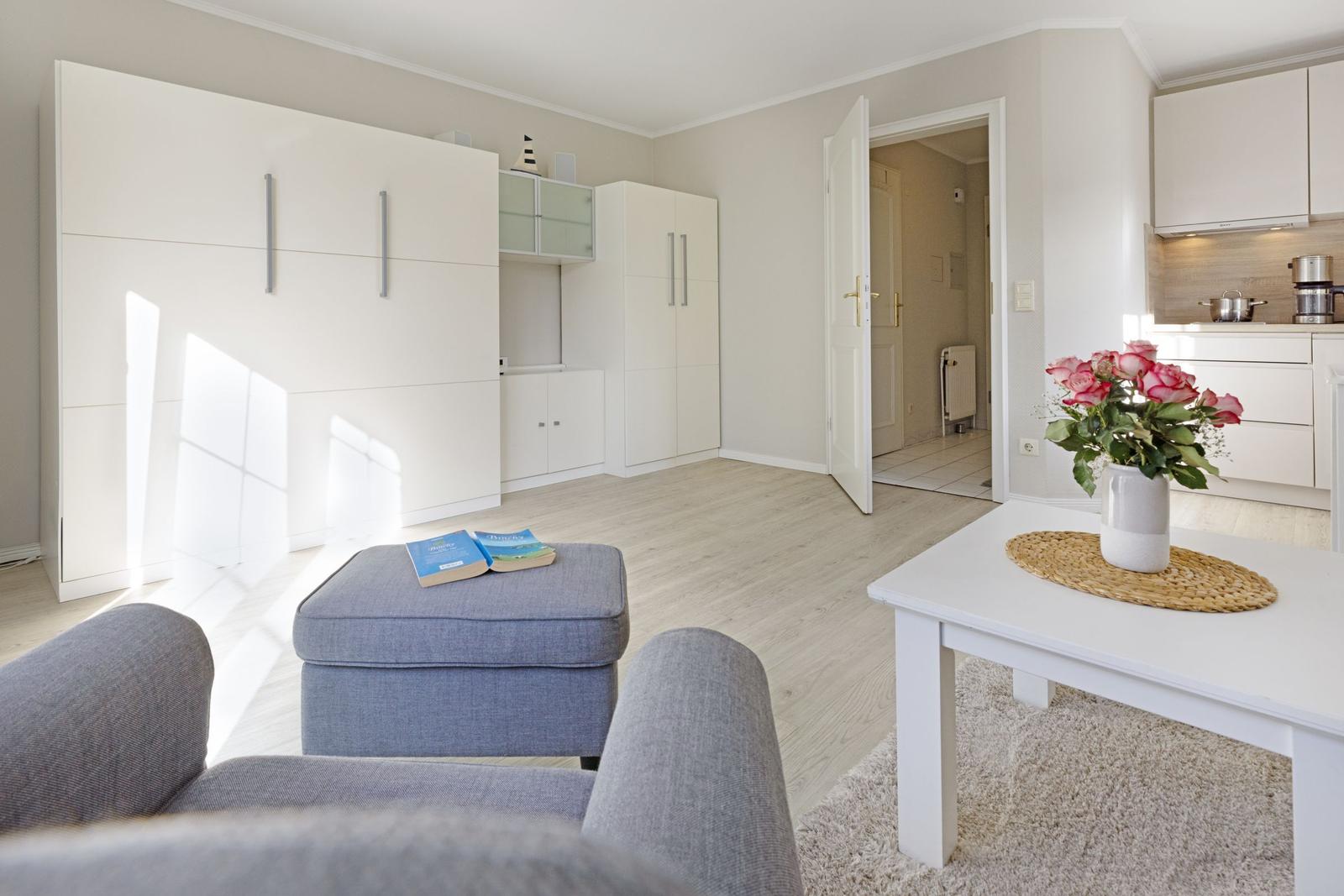 Living area with white cabinets, gray sofa, and kitchen area with flowers.