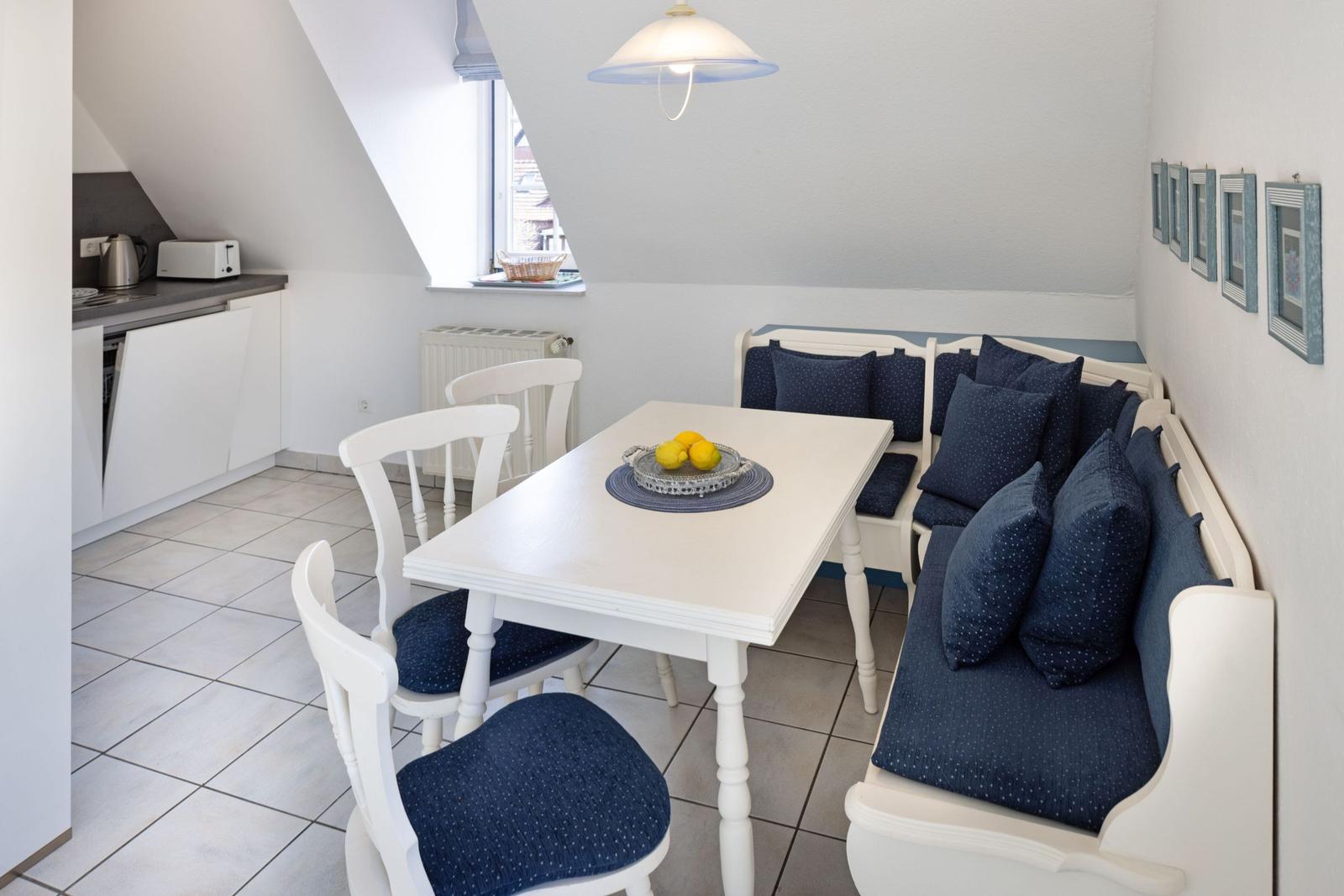 Esszimmer mit weißem Tisch, blauen Sitzkissen und Küchenbereich im Hintergrund.