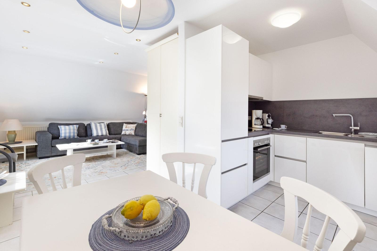Küche mit Essbereich und Wohnzimmer im Hintergrund. Weiße Möbel und graue Wand.