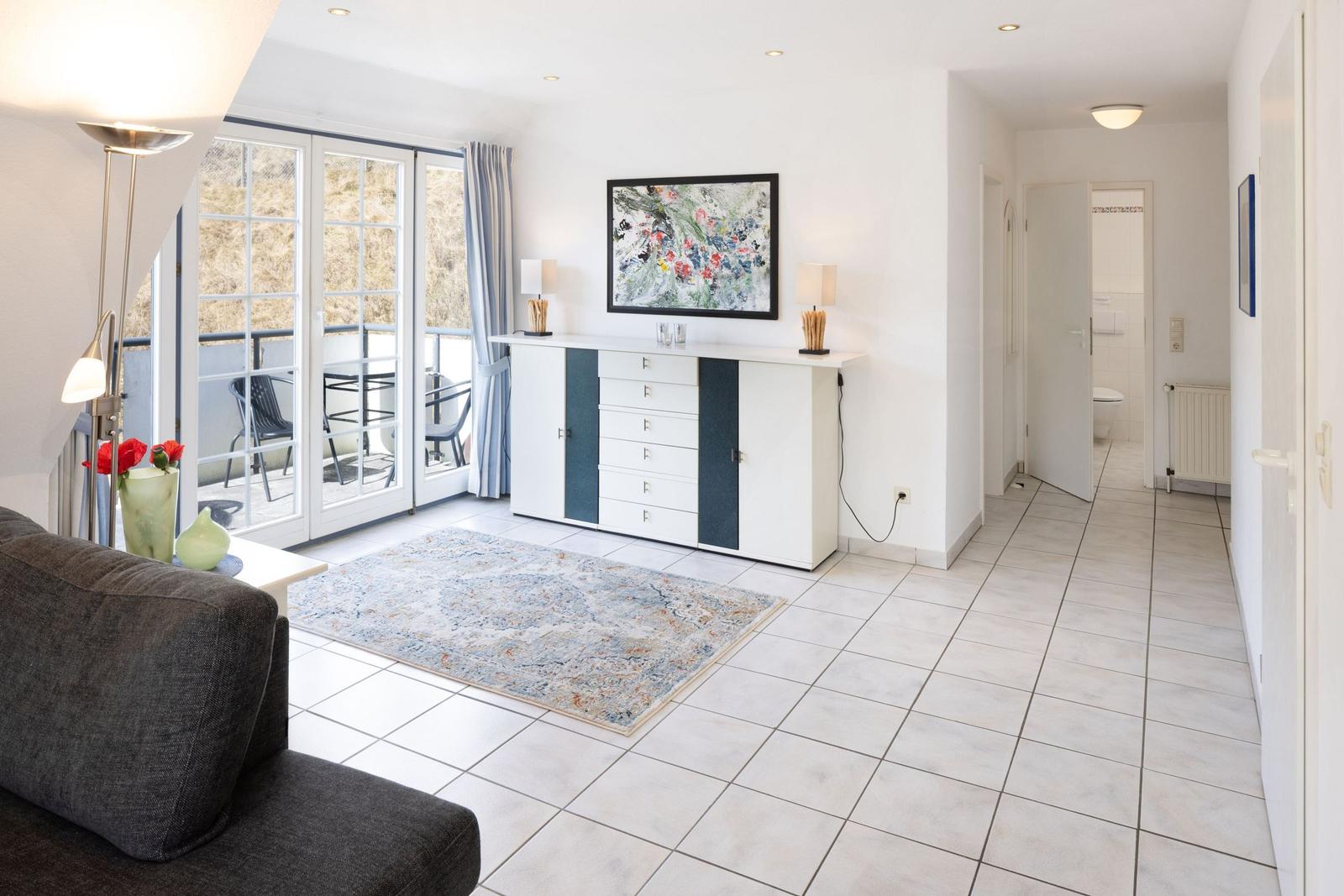 Wohnzimmer mit Schlafzimmer und Bad im Hintergrund. Fliesenboden und Balkon mit Sitzgelegenheiten.