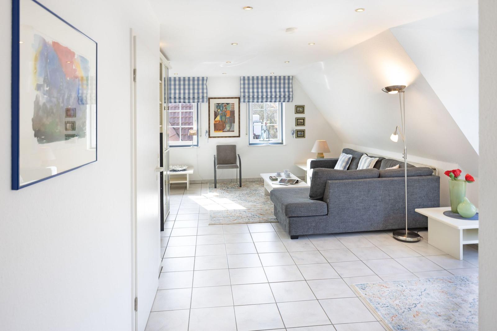 Wohnzimmer mit grauem Sofa, weißen Wänden und Kachelboden. Fenster mit karierten Rollläden.
