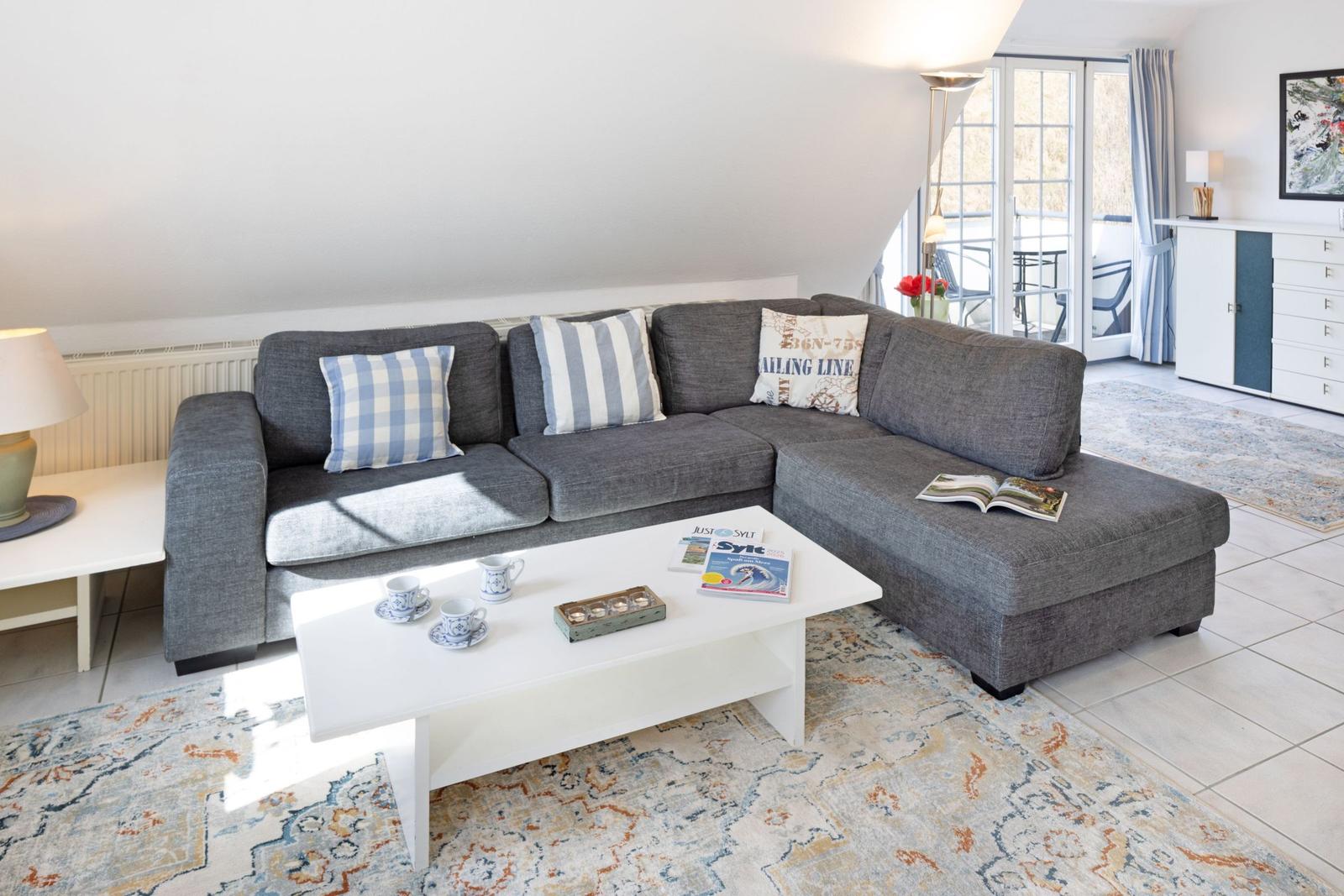 Ein Wohnzimmer mit grauem Sofa, weißem Couchtisch und Teppich. Hinter dem Sofa sind Fenster mit Blick auf Balkon.
