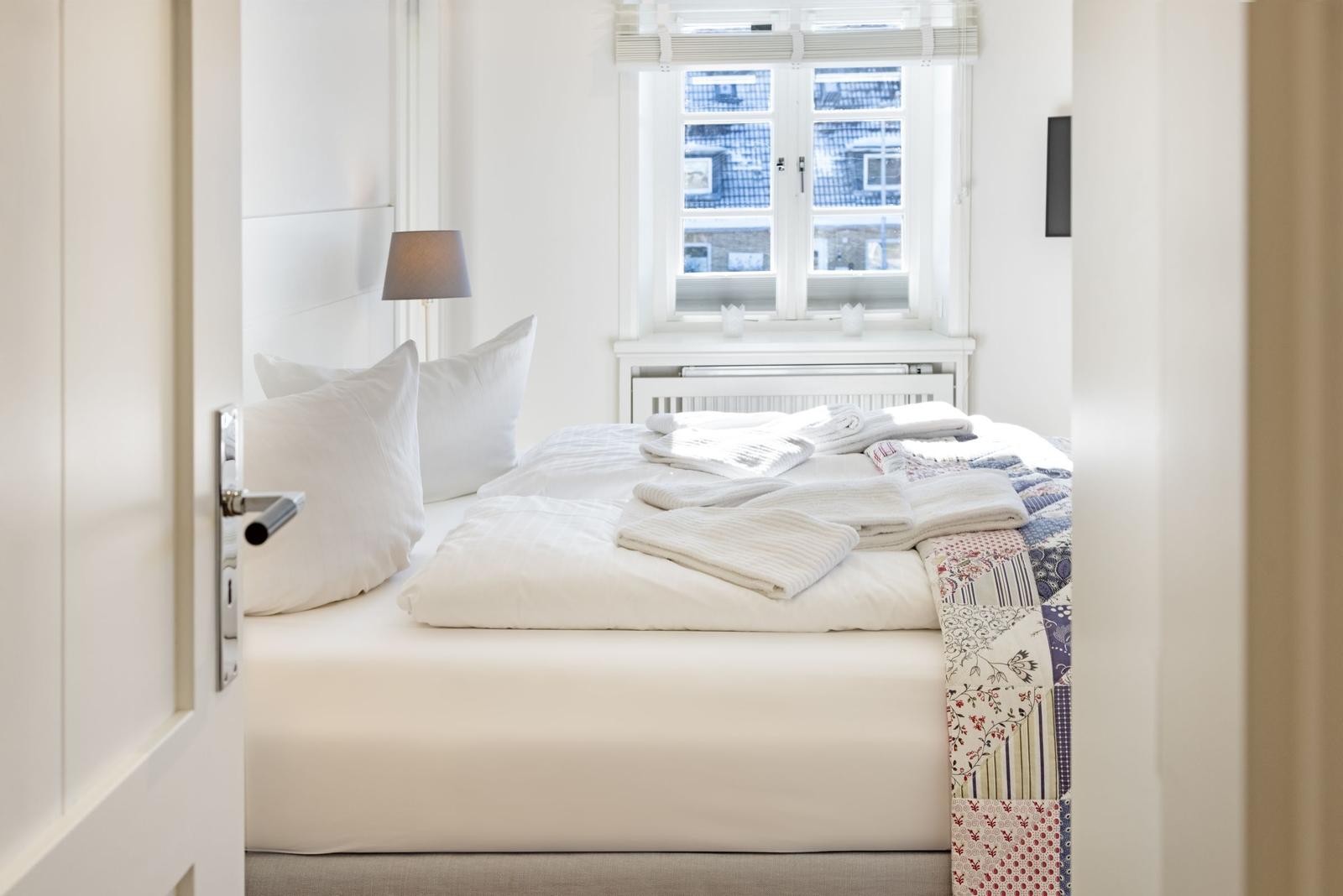 Ein Schlafzimmer mit weißem Bett, Kissen und einem Fenster mit Blick auf Dächer.