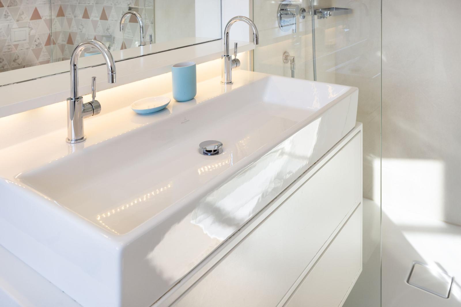 Waschbecken mit zwei Wasserhähnen und Spiegelschrank. Dusche mit Glaswand daneben.