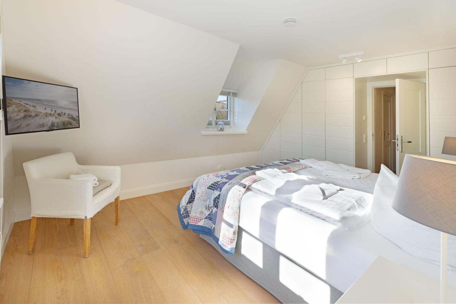 Schlafzimmer mit Dachschräge, Bett, Stuhl und Fernseher an der Wand.