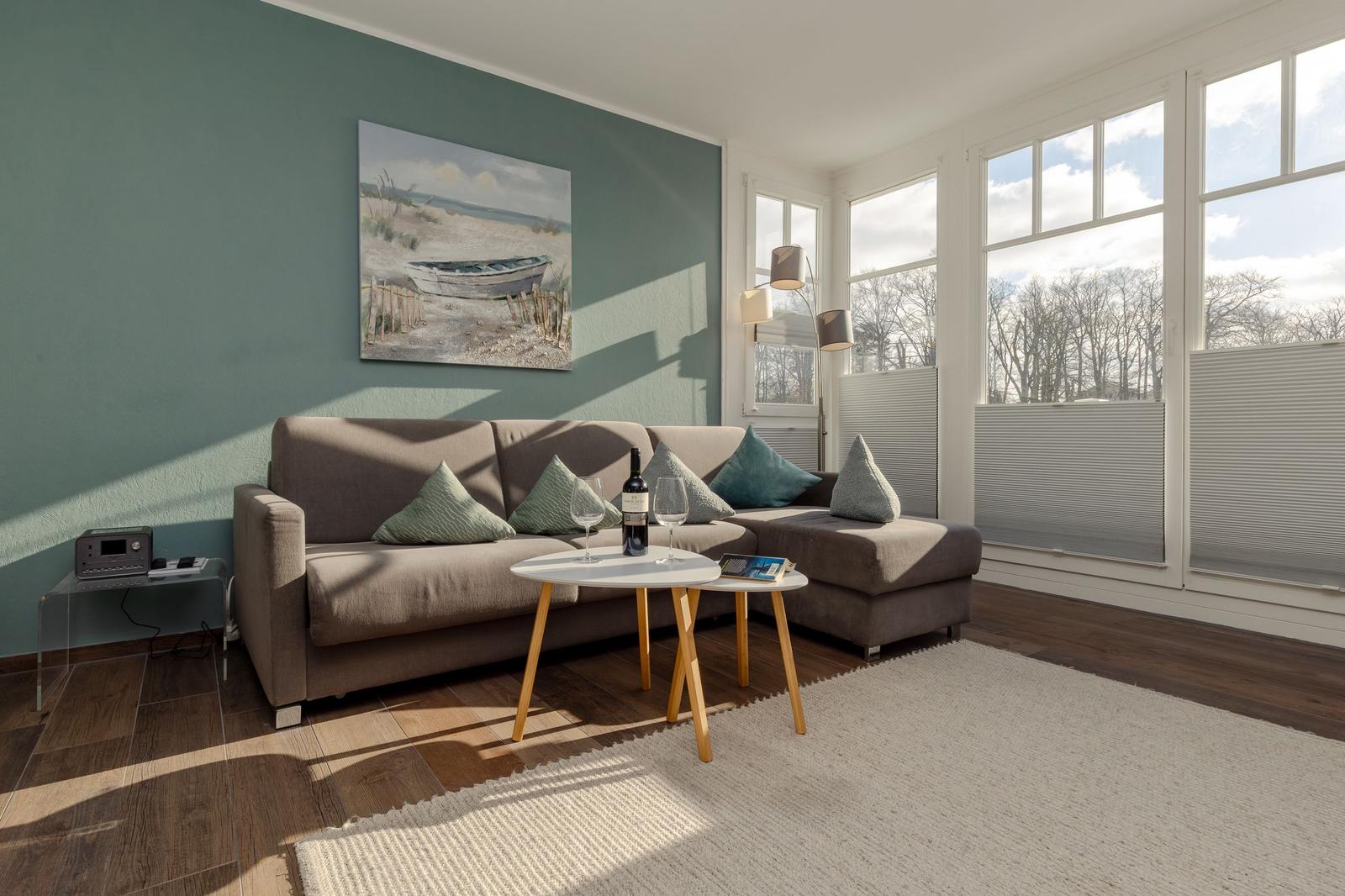 Wohnzimmer mit Sofa, Tisch und großen Fenstern. Wandbild zeigt Boot.
