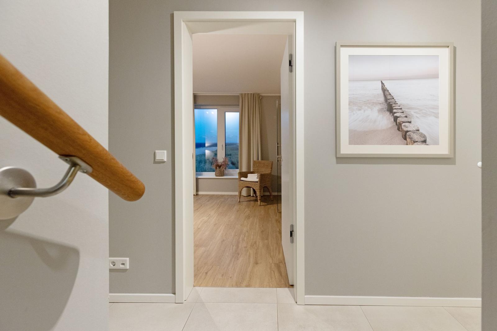 Hausgang mit Holzgeländer und Bild an der Wand. Durch Tür ist Wohnzimmer mit Fenster und Stuhl sichtbar.