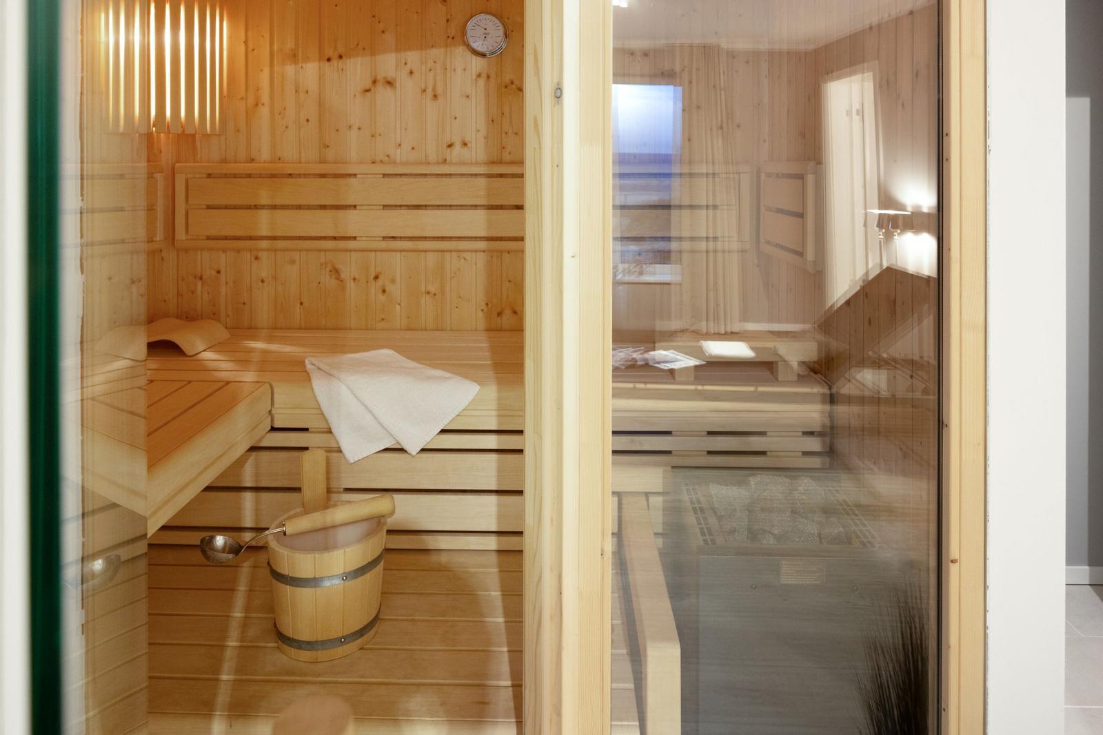 Sauna mit Holzbank, Körbchen und Thermometer. Glaswand zeigt Badebereich.