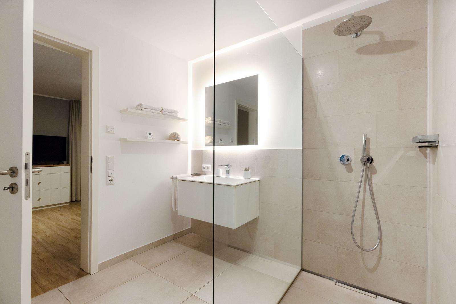 Badezimmer mit Dusche, Waschbecken und Spiegel. Durchgang zur Wohnung.