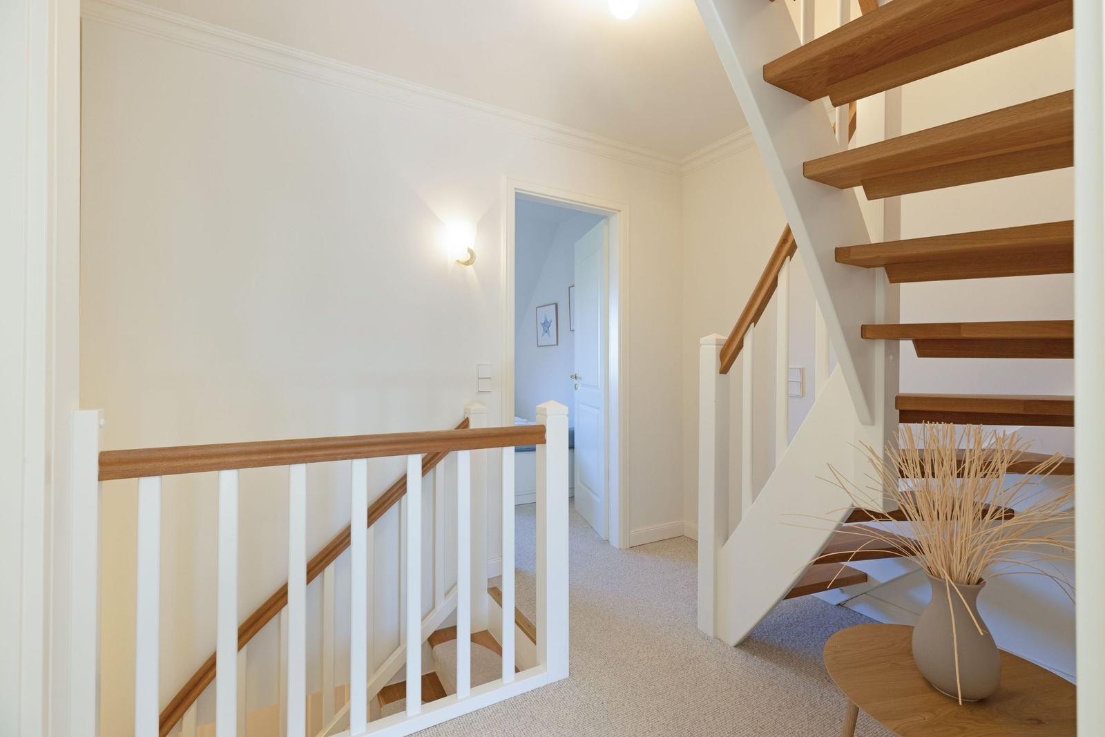 Treppenhaus mit Holzstufen und weißer Balustrade. Im Hintergrund ist ein Flur mit Tür zu sehen.