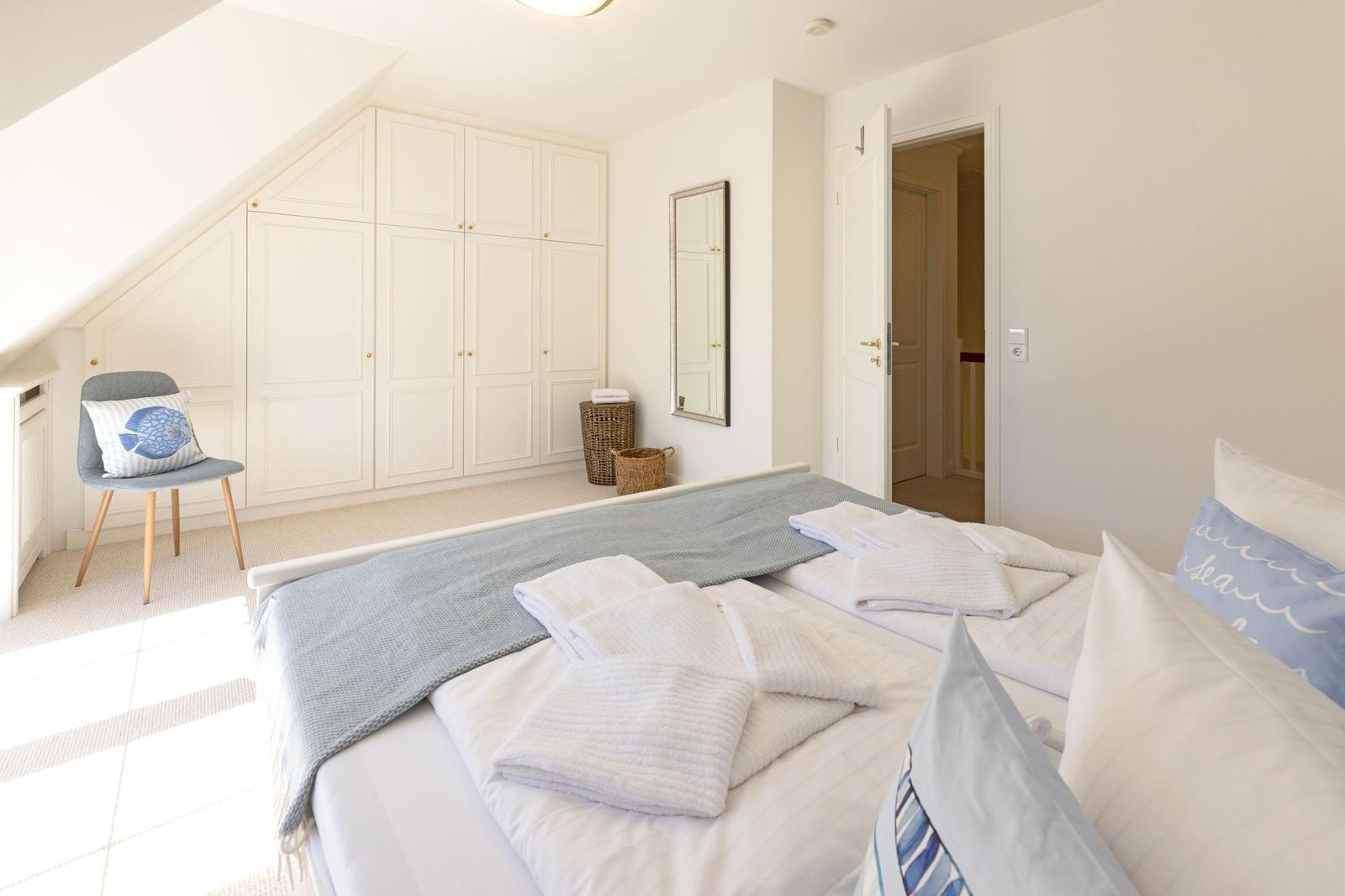 Schlafzimmer mit Dachschrägen, weißer Einbaukombination und Bett mit weißen Bettwäsche und blauen Kissen.