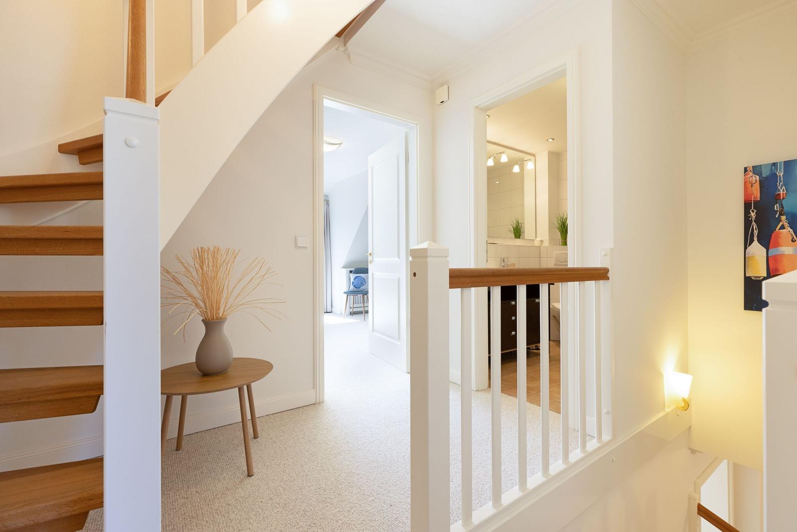 Hausflur mit Holztreppe, kleinem Tisch und Vasen. Blick in Schlafzimmer und Bad.