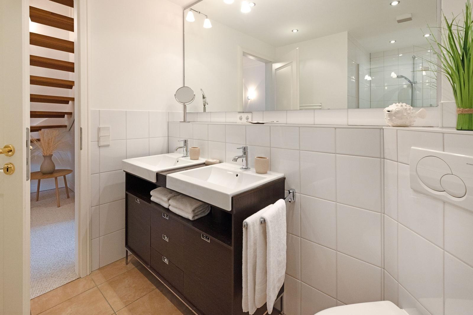 Badezimmer mit Doppelwaschbecken, Spiegel und Toilette. Holztreppe im Hintergrund.