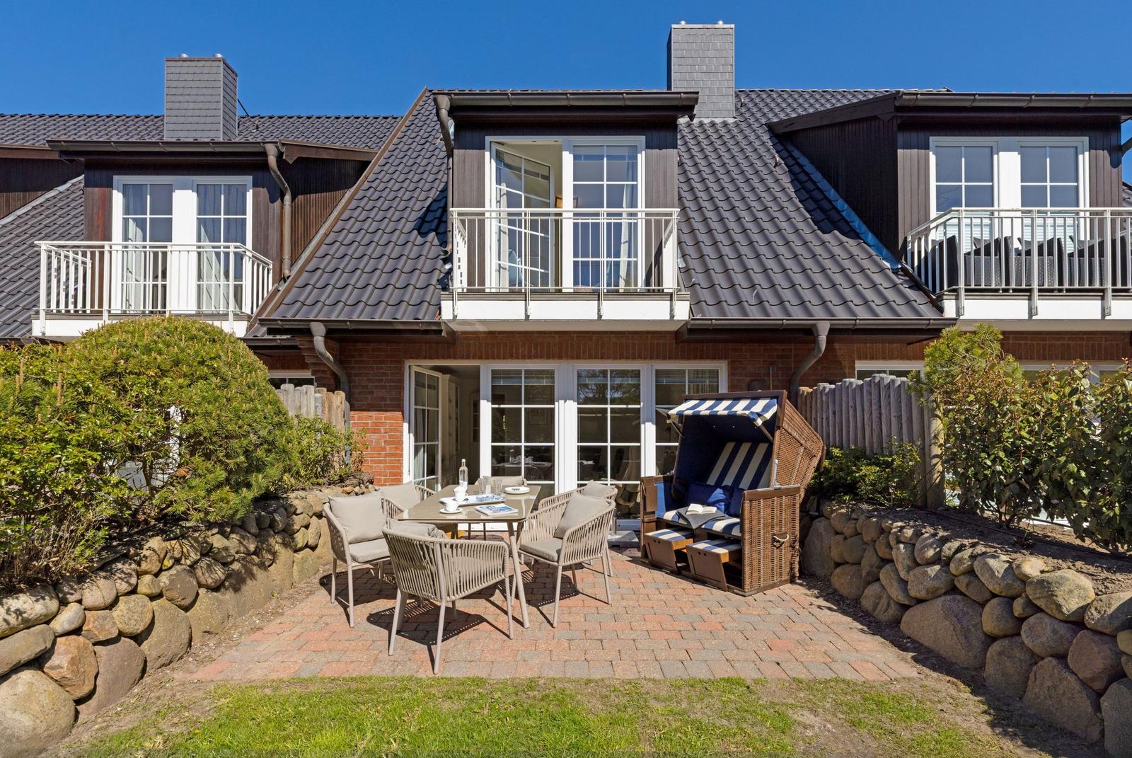 Terrasse mit Tisch, Stühlen und Strandkorb. Hintergrund: Haus mit Balkon und Dach.