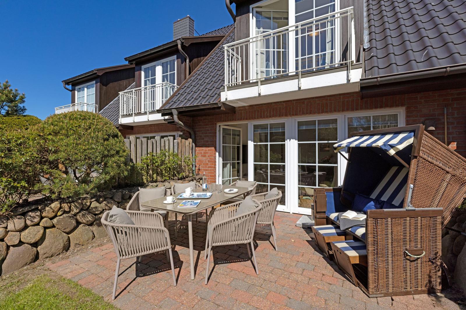 Terrasse mit Tisch, Stühlen und Strandkorb. Hintergrund: Haus mit Balkon und Glasfront.