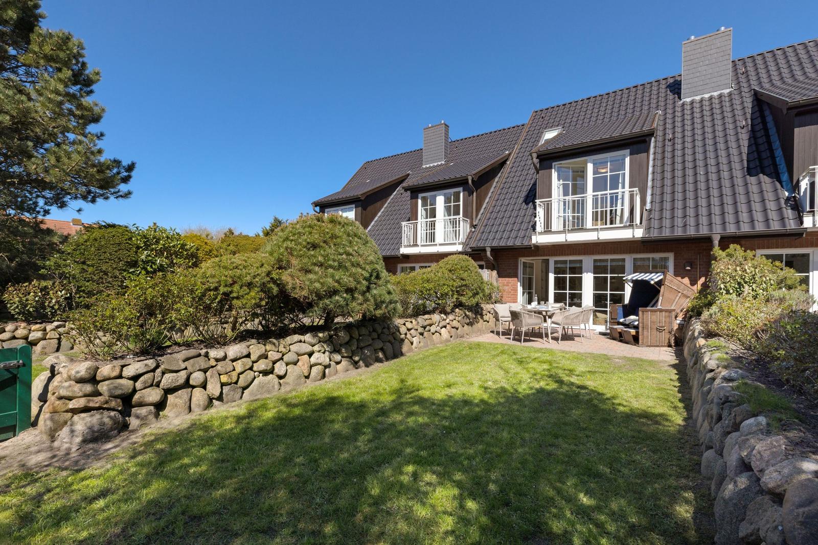 Haus mit Balkon, Terrasse und Garten. Steinmauer und Bäume umgeben den Außenbereich.