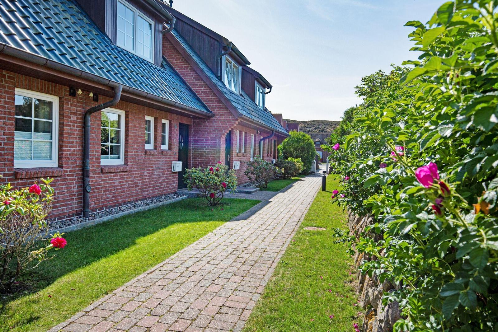 Ein gepflasterter Weg führt entlang roter Backsteinhäuser mit grünen Gärten und blühenden Rosen.