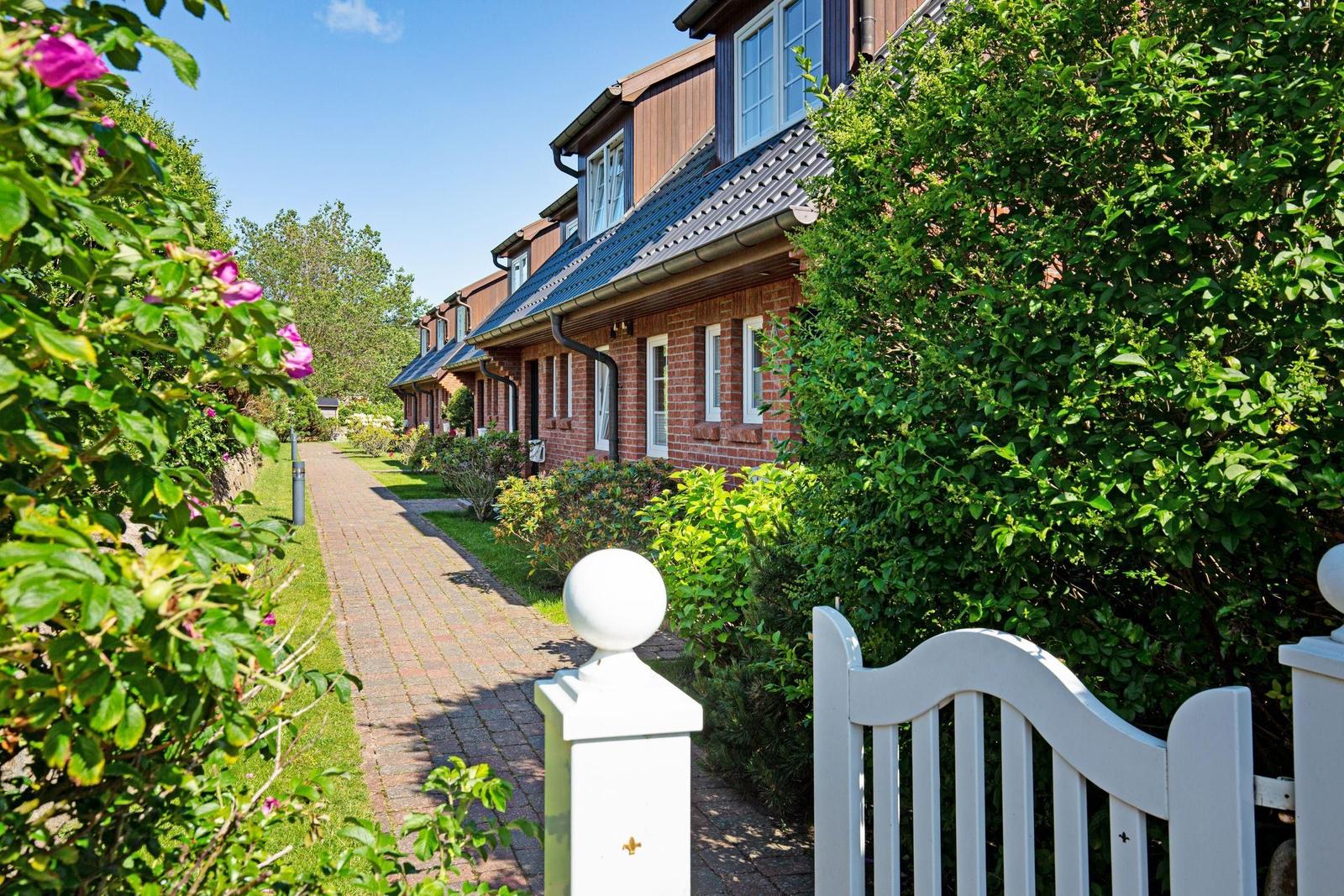 Ein gepflasterter Weg führt an roten Backsteinhäusern vorbei, umgeben von grünen Sträuchern und Blumen.