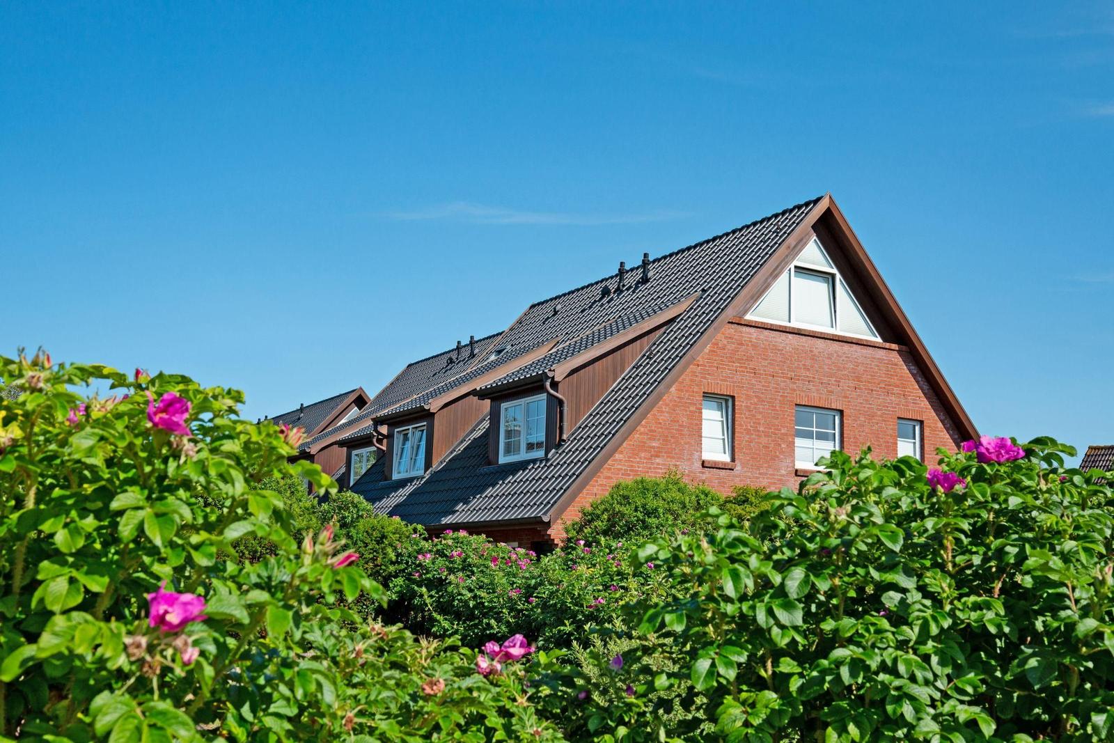Ein Haus mit rotem Backstein und dunklem Dach, umgeben von grünen Sträuchern und rosa Blüten.