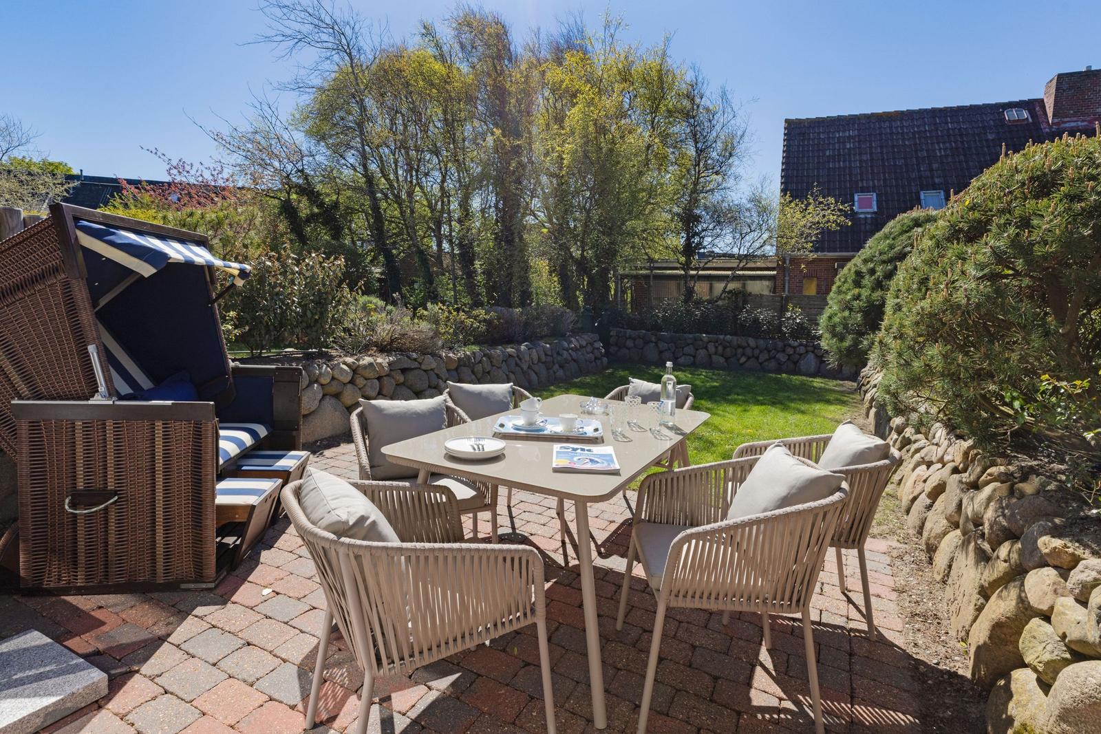Terrasse mit Tisch, Stühlen und Strandkorb. Grünfläche und Steinmauer im Hintergrund.