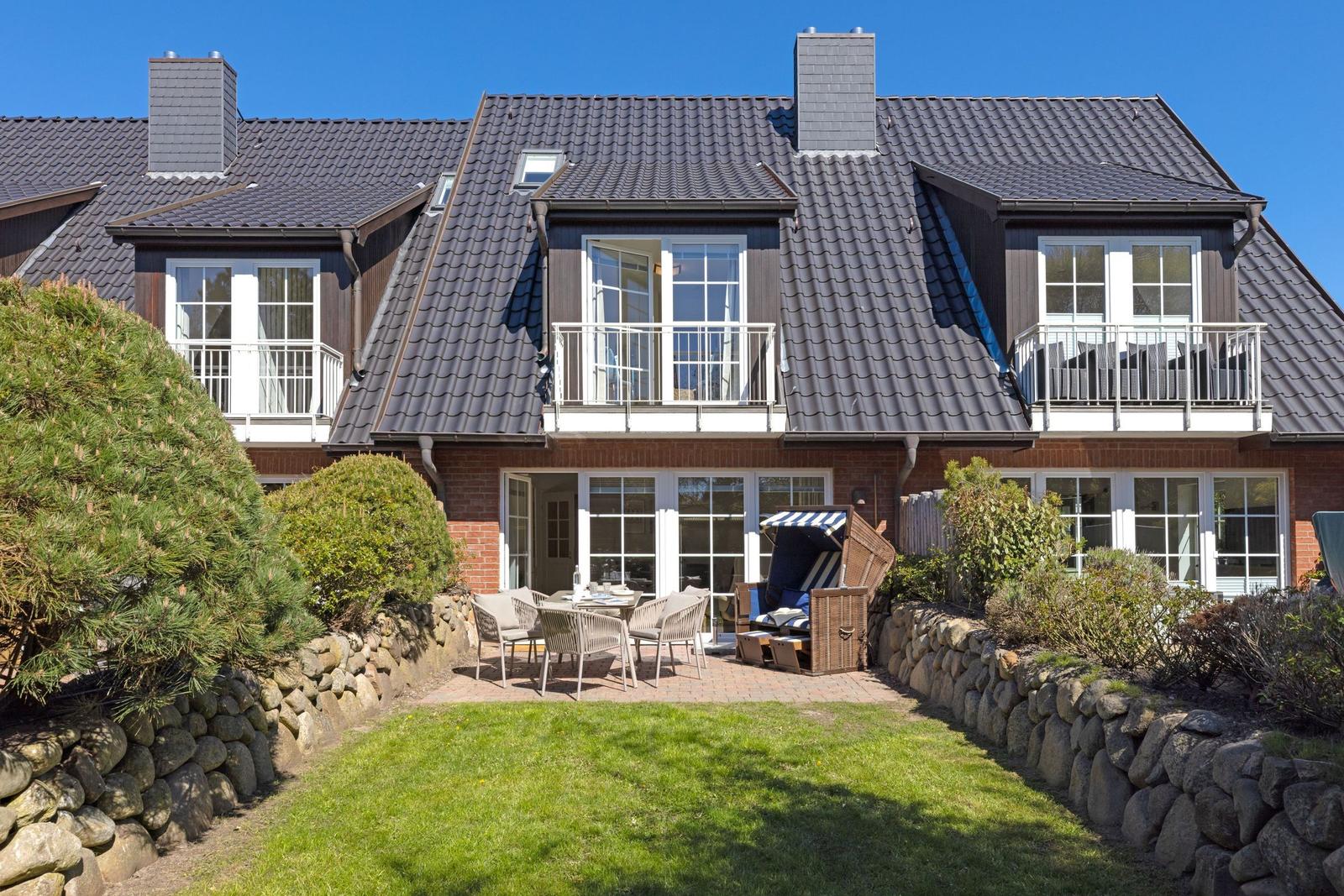 Haus mit Balkon, Terrasse und Gartenbereich. Holz- und Steinmauern umgeben den Außenbereich.