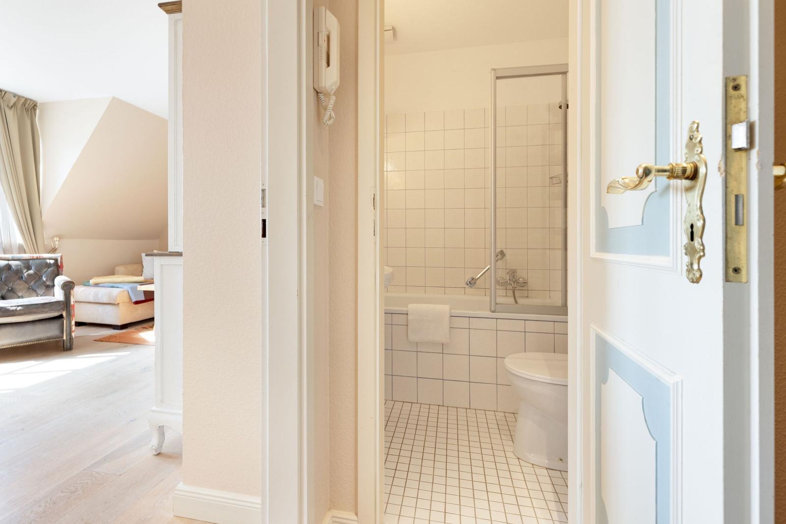 Badezimmer mit Badewanne und Dusche. Blick in Schlafzimmer mit Sofa und Fenster.