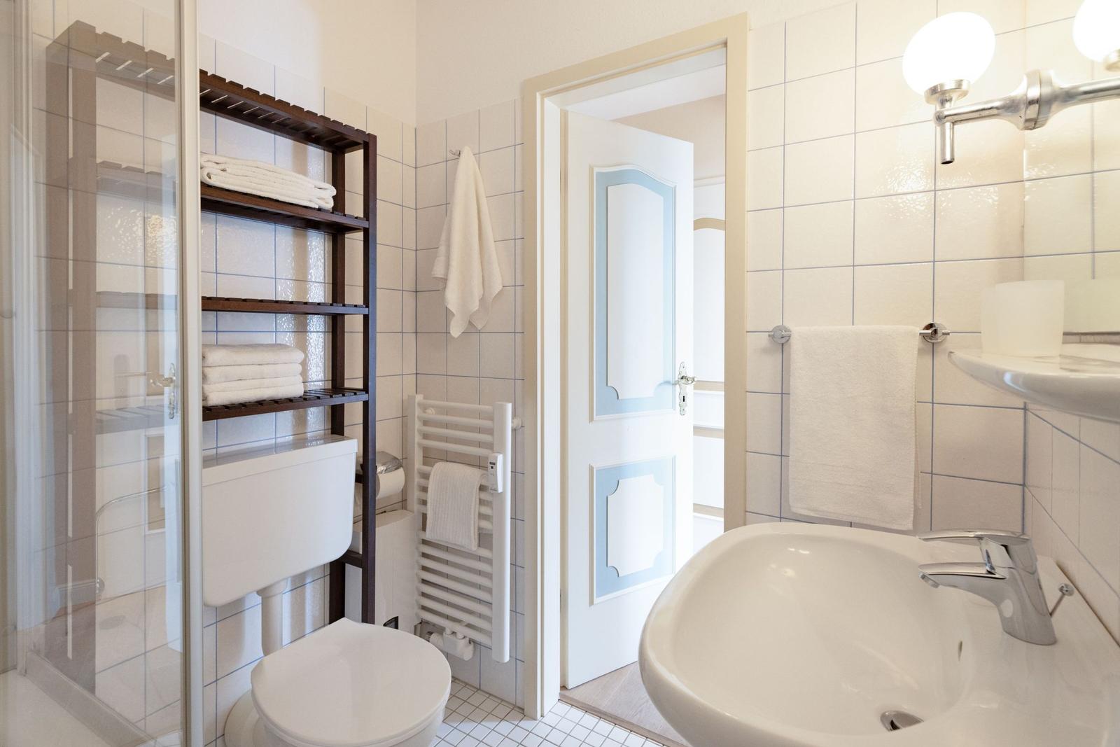 Badezimmer mit Dusche, Waschbecken und Toilette. Weiße Fliesen und Handtücher.