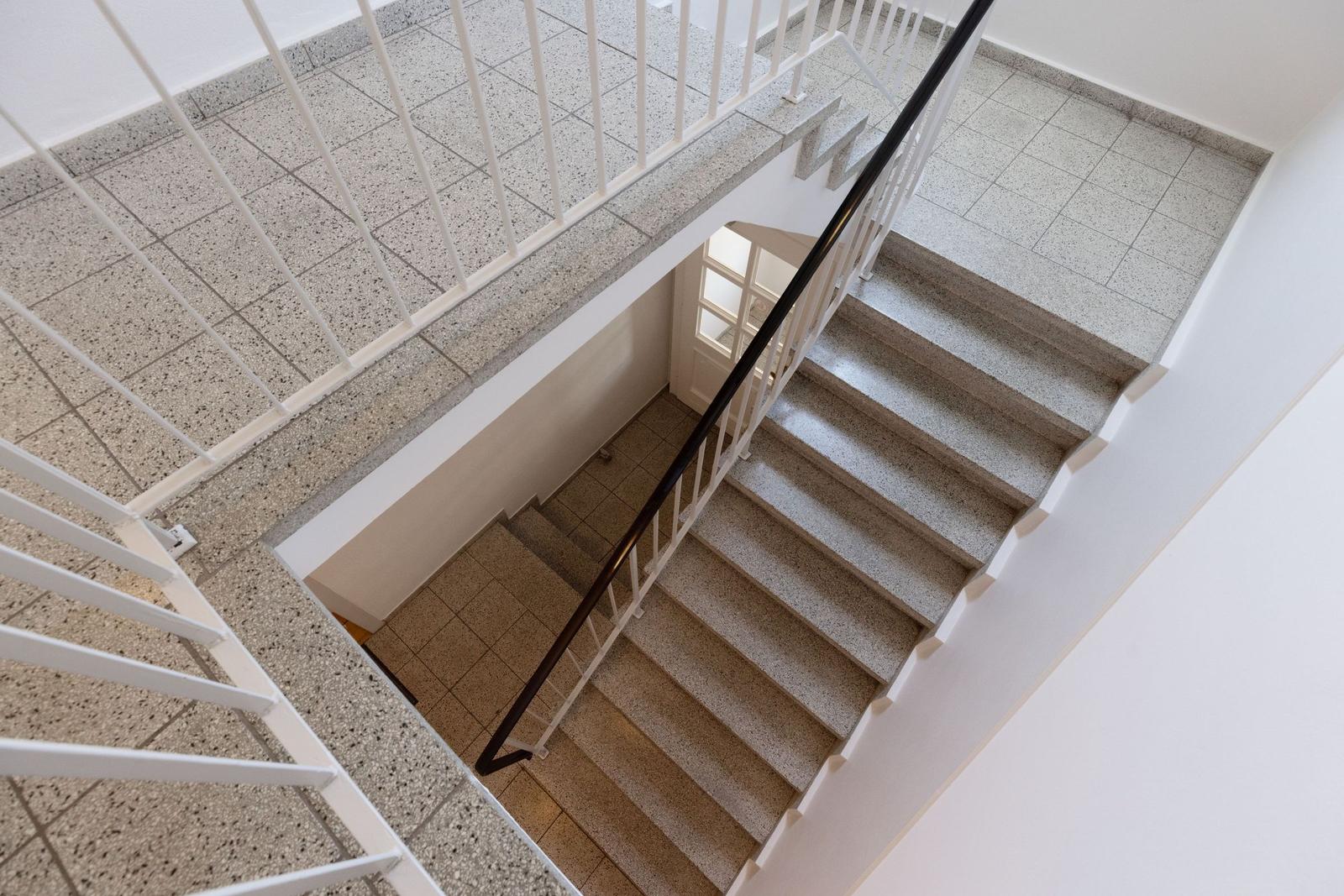Treppe mit weißem Geländer und dunklem Handlauf. Bodenfliesen und weiße Wände.