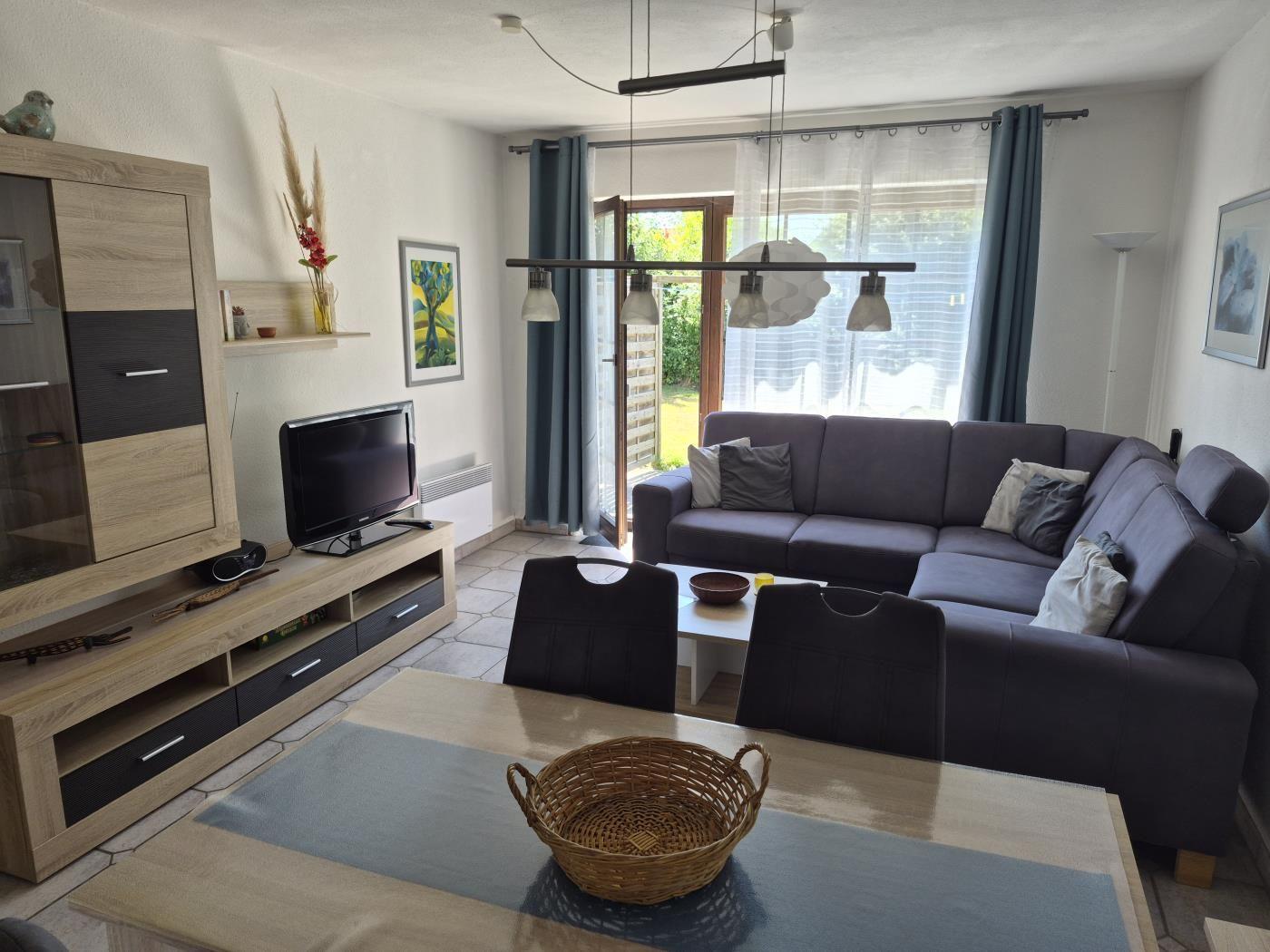 Wohn- und Essbereich mit Sofa, Fernseher und Tisch. Durchgang zur Terrasse.