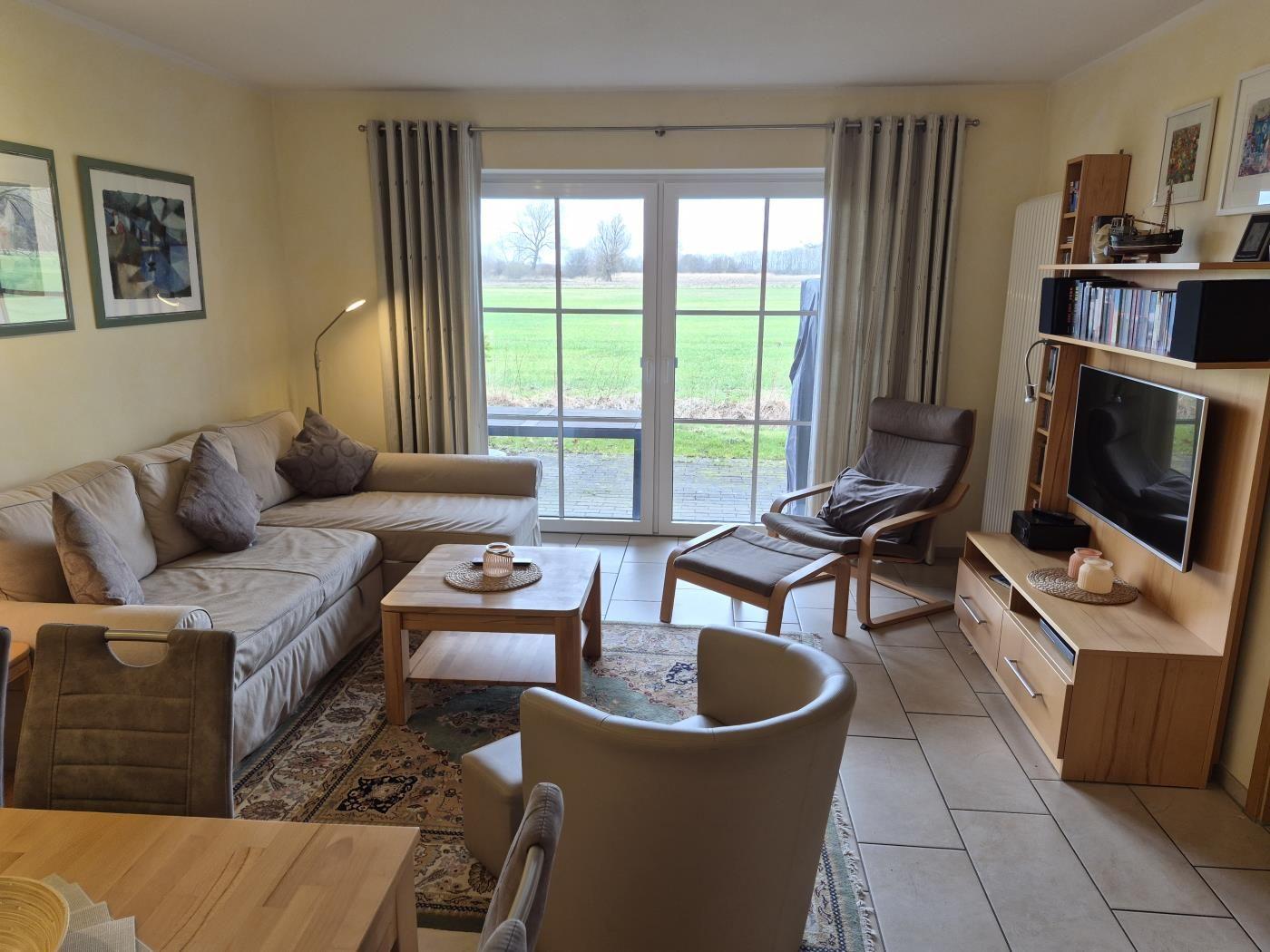 Wohnzimmer mit Sofa, Sessel und Fernseher. Durchgangstür führt auf eine Terrasse mit Blick auf Feld.