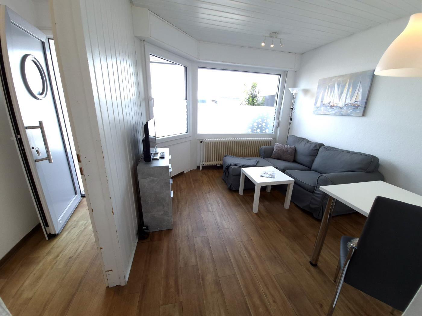 Wohnbereich mit Sofa, Tisch und Fenstern. Holzboden und Wandbild mit Segelbooten.