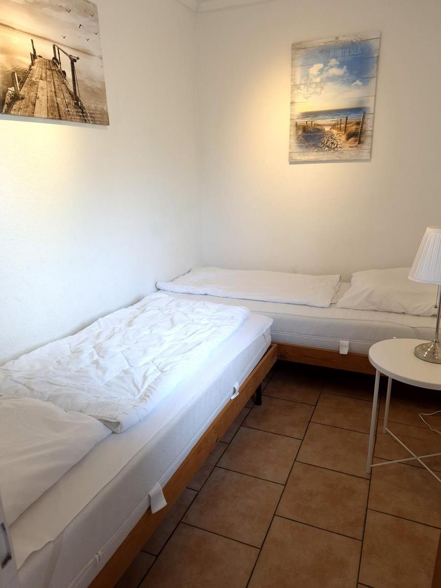 Zimmer mit zwei Betten, Wandbildern und Tischlampe auf Fliesenboden.