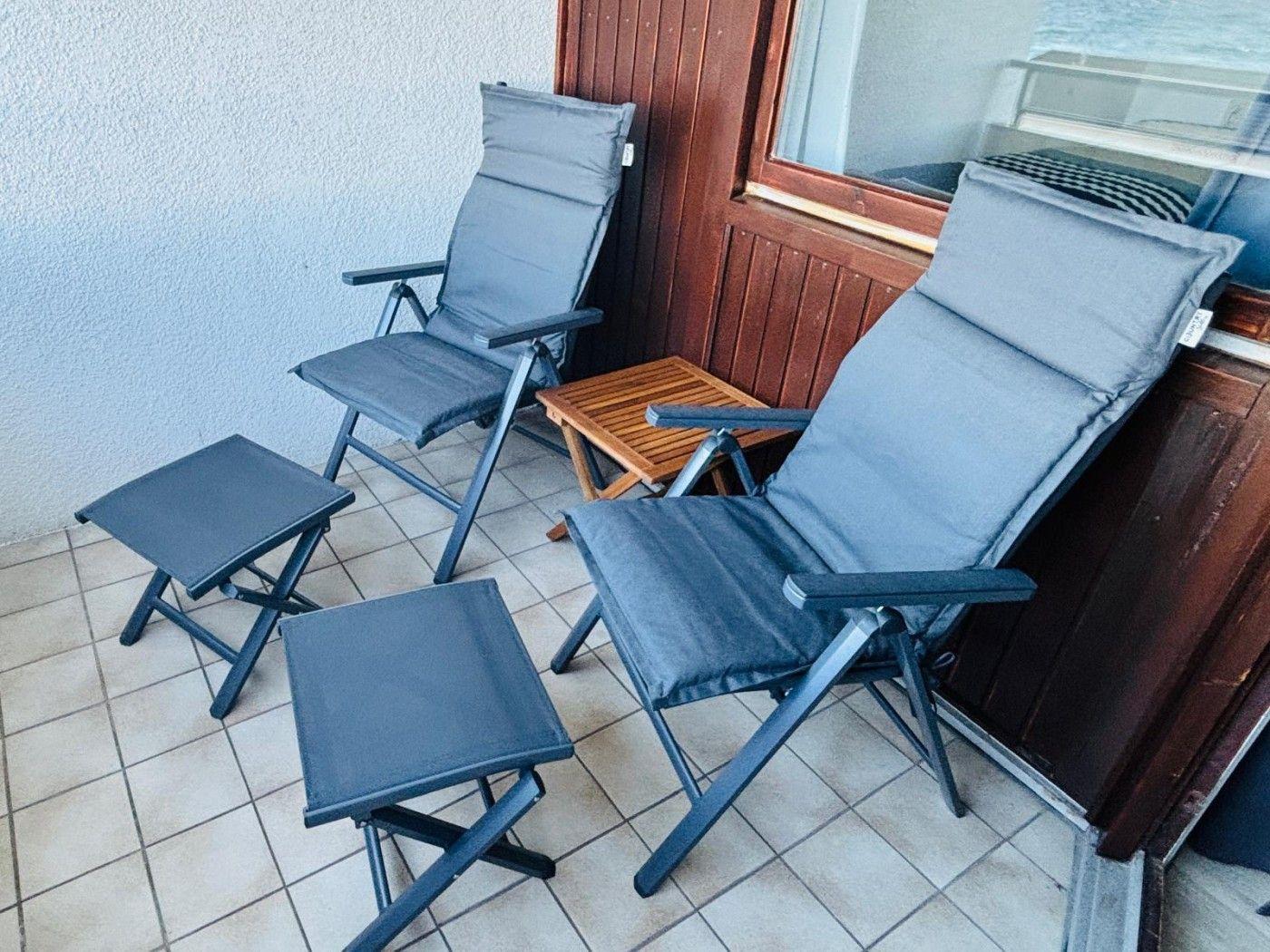 Terrasse mit zwei Liegestühlen und zwei Fußstützen. Holztisch und Holzwand sichtbar.